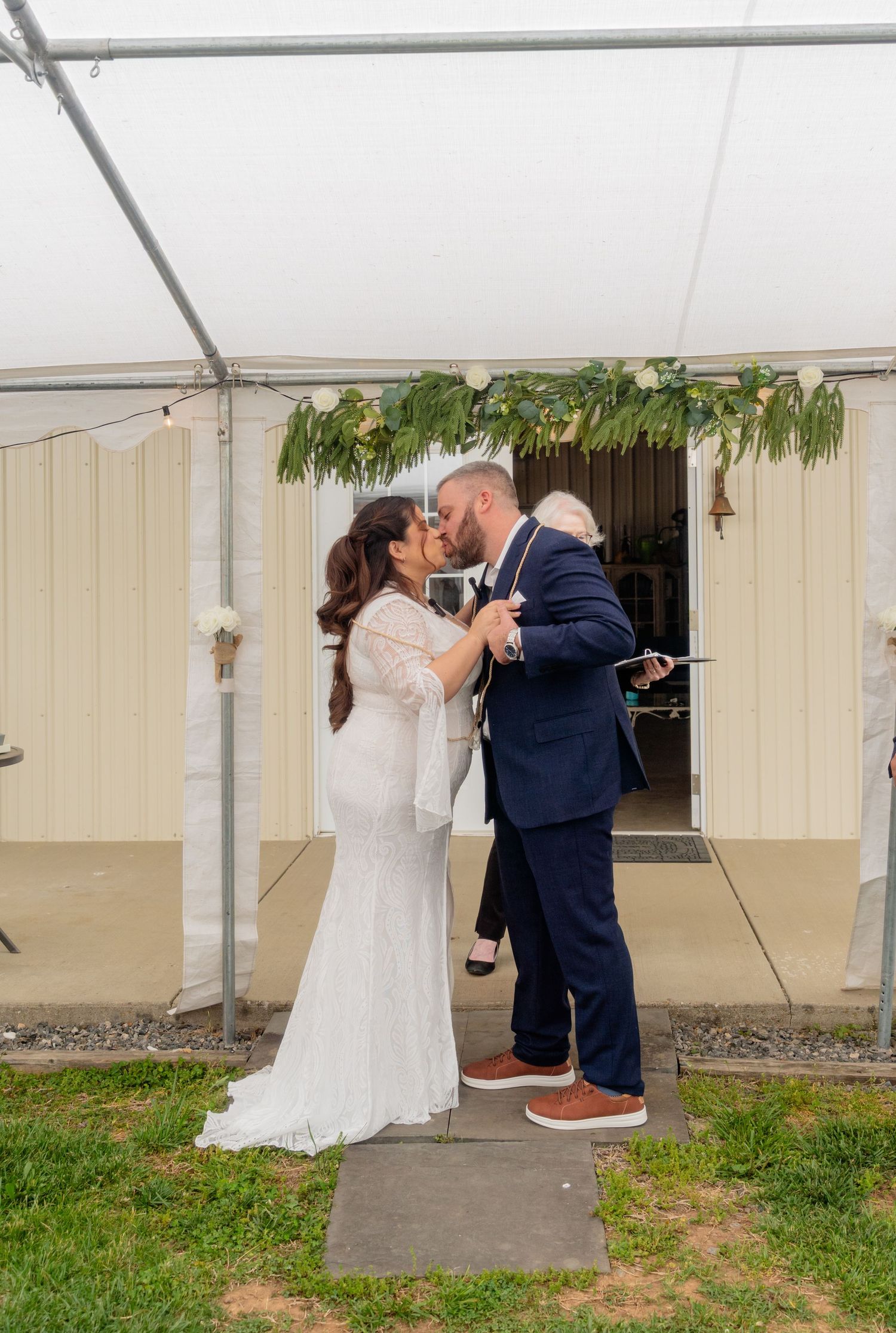 Bride and groom share first kiss under a white tent adorned with greenery at an outdoor wedding ceremony.