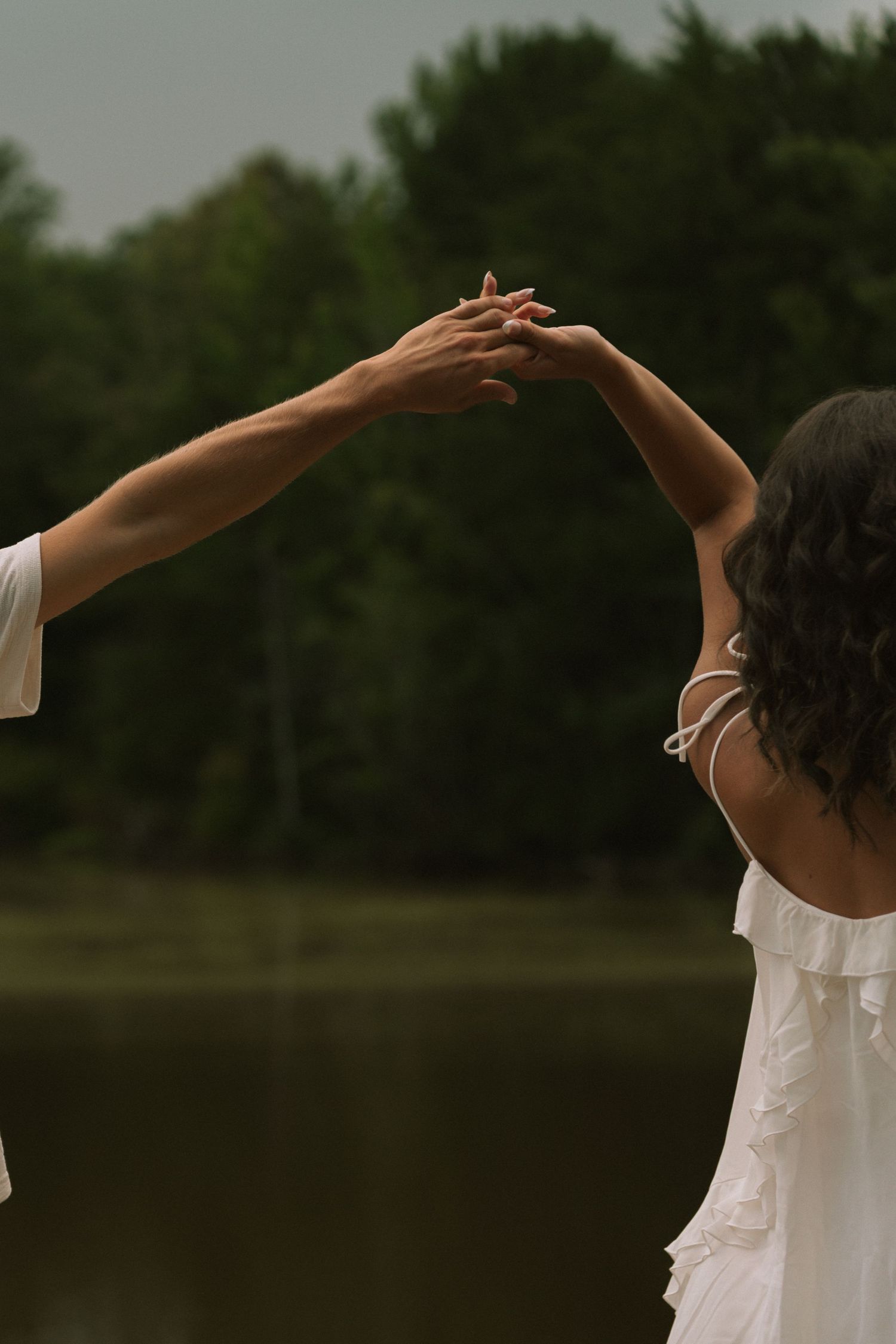 Two people in white dresses hold hands raised above their heads near a serene lake surrounded by lush green trees.