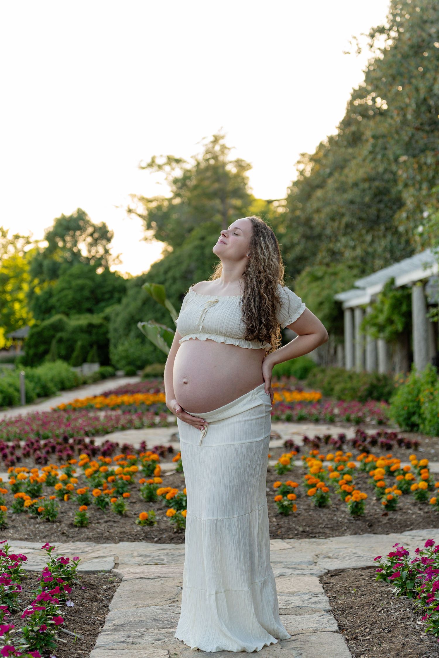 Pregnant woman in white dress cradling baby bump in a colorful garden at golden hour. Taken by Row And Co Photography in Maymont RVA