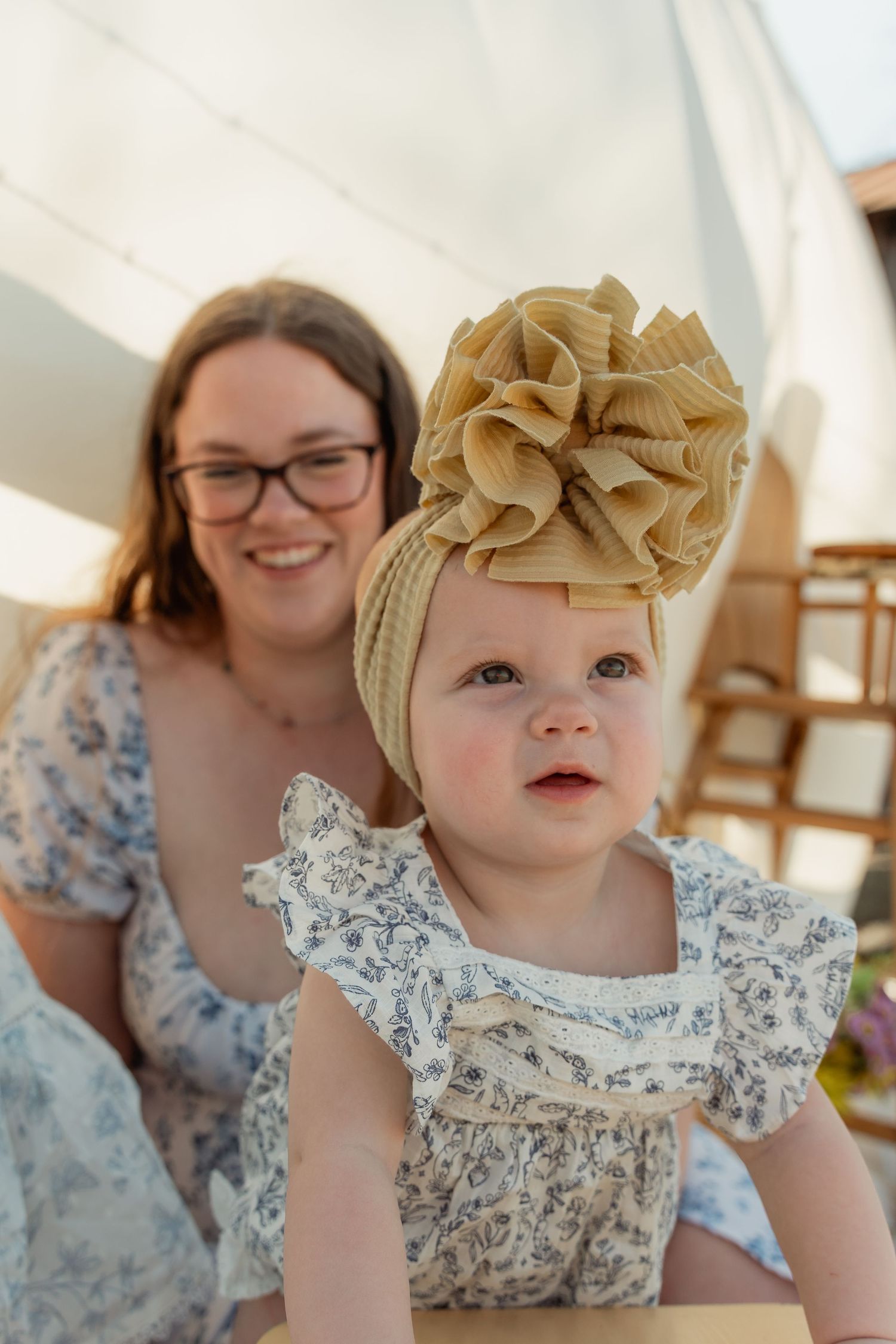 Adorable baby girl in floral dress wearing a large bow turban headband with smiling mother behind. Taken by Row And Co Photography in FXBG