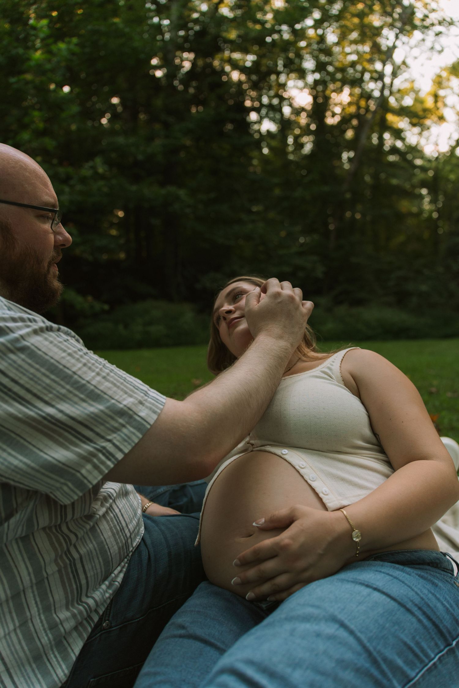 A pregnant woman smiles as her partner tenderly touches her face while she holds her baby bump outdoors.