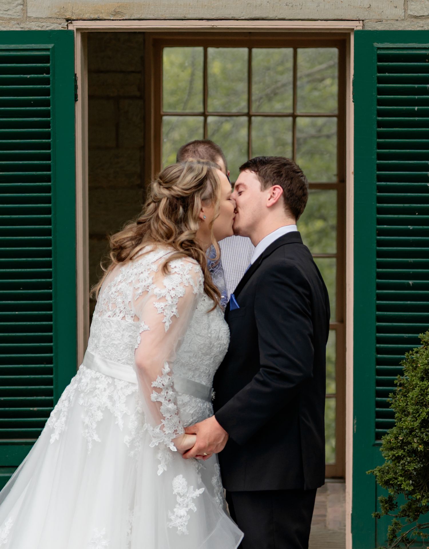 Bride and groom sharing a first kiss in wedding attire outside a stone building with green shutters.