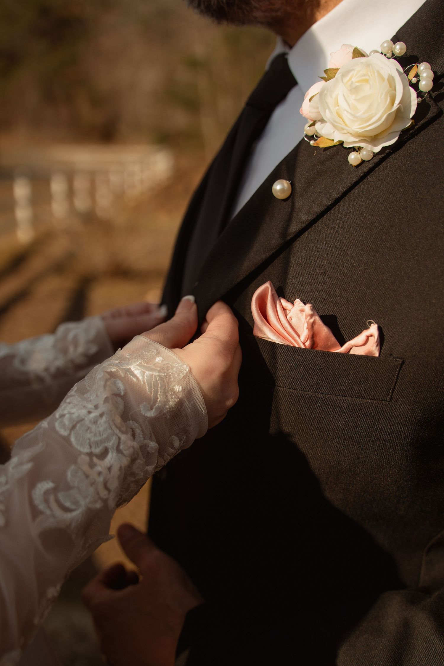 Bride adjusting groom's dark suit boutonniere with white rose and pearl accents, pink pocket square visible.