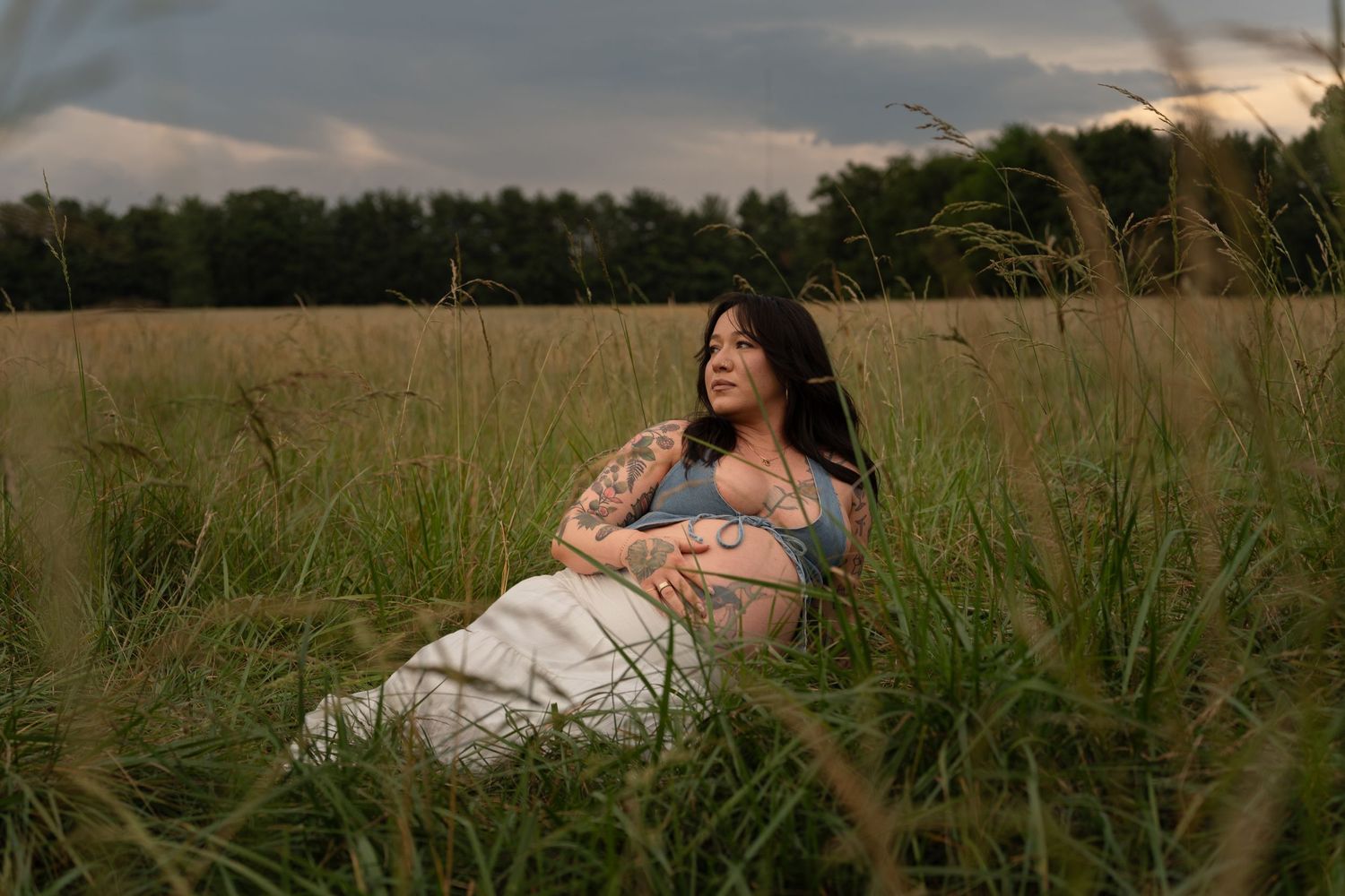 Pregnant woman with tattoos sits peacefully in tall grass field at dusk, wearing white skirt and blue crop top.