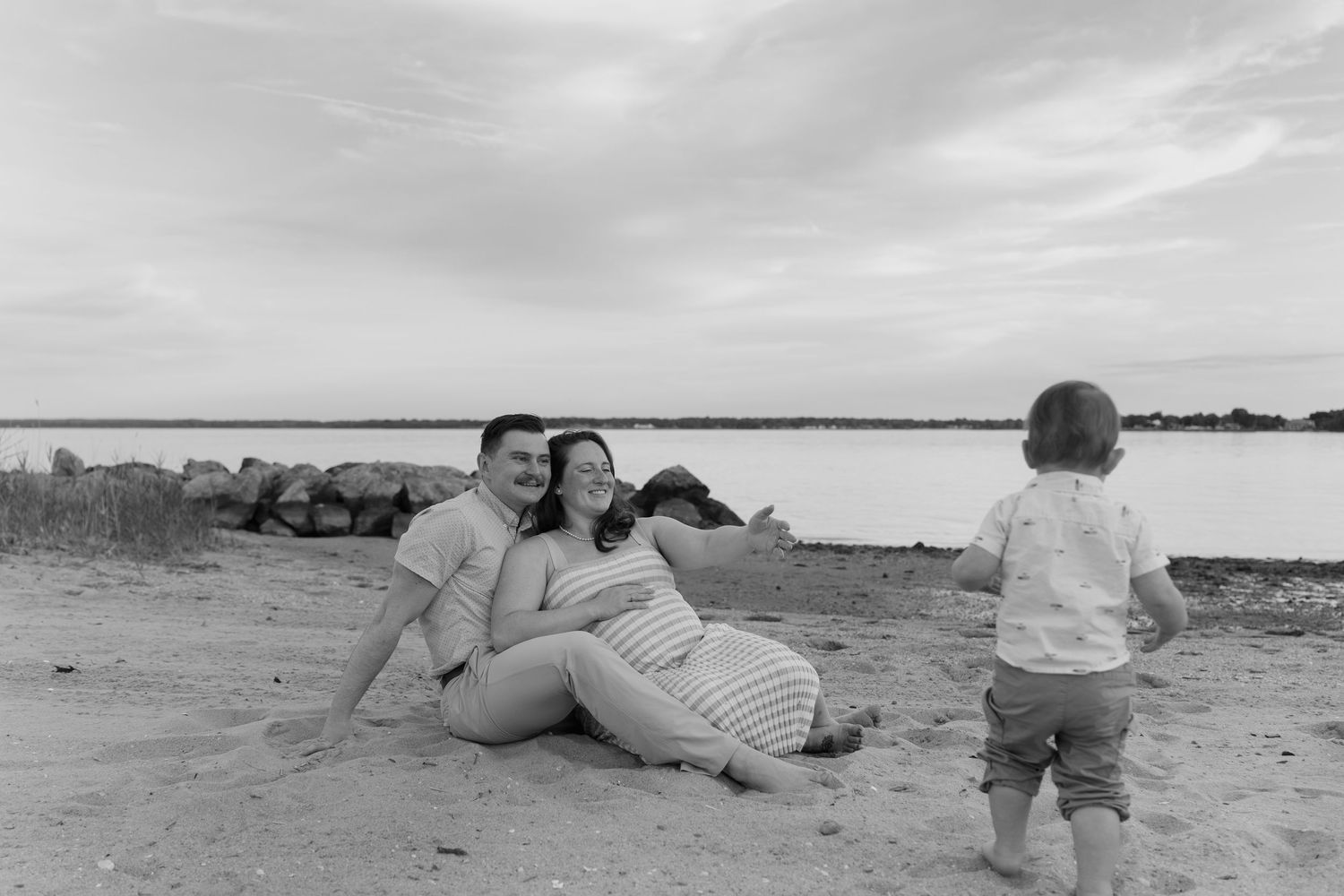 Black and white beach photo of a pregnant couple sitting on sand smiling at a toddler standing nearby.