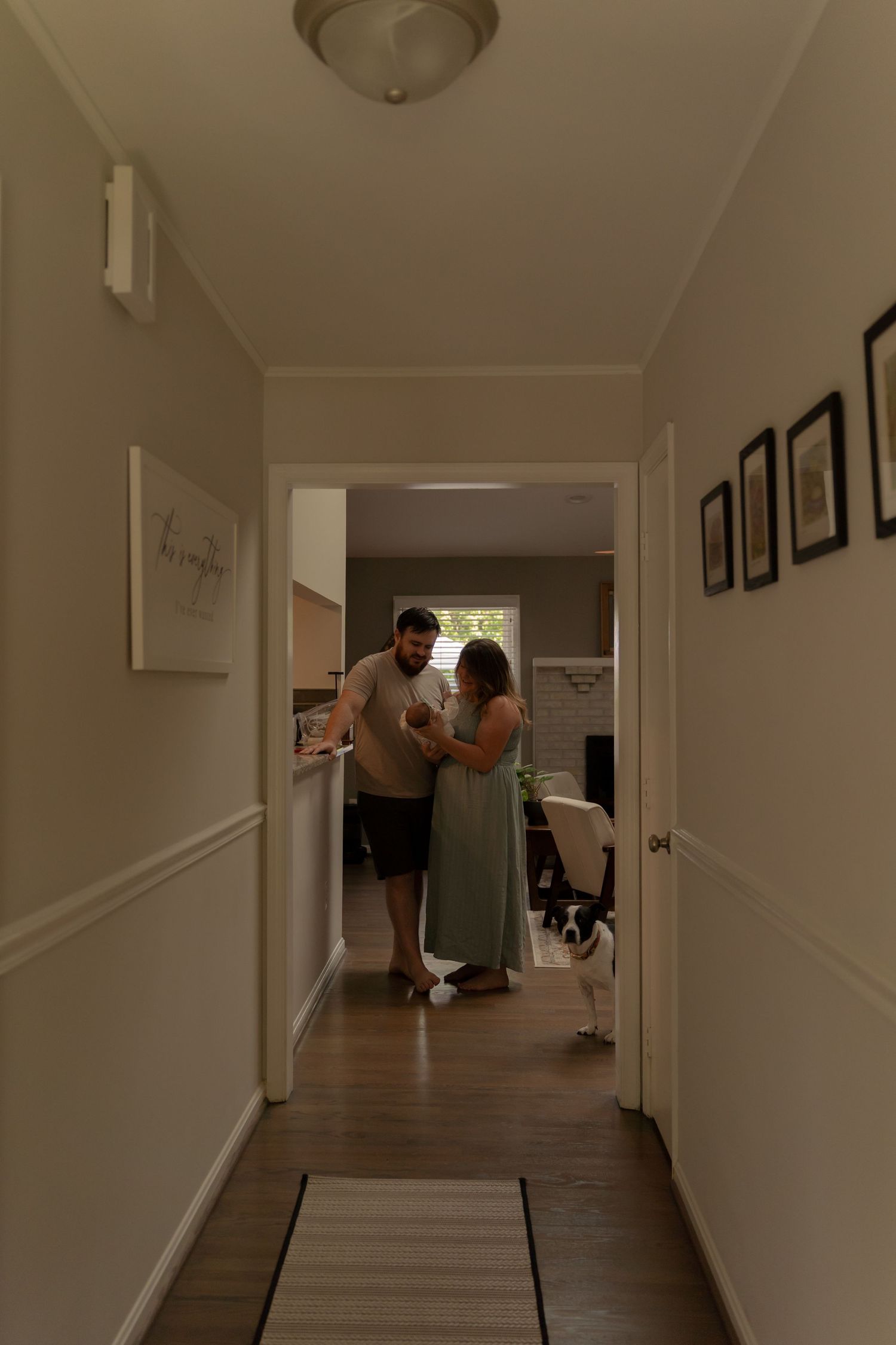 A couple stands in a warmly lit hallway holding a newborn baby while a dog watches nearby in their cozy home.