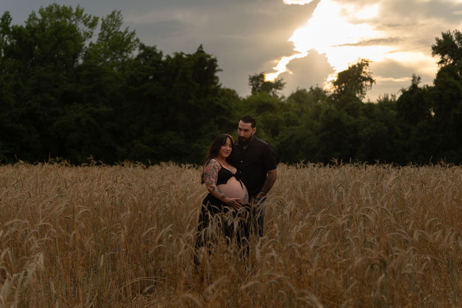 Couple embracing in golden wheat field at sunset with dramatic clouds and lush green trees in background.