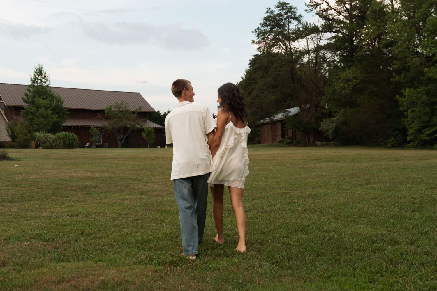 Couple walking hand in hand across a green lawn near a rustic building surrounded by trees at dusk.
