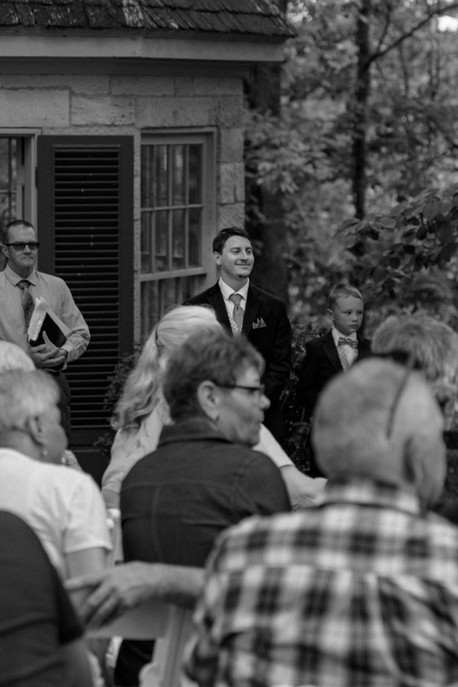 Black and white outdoor wedding ceremony with smiling groom in suit standing before seated guests near rustic building. Row and Co Photo