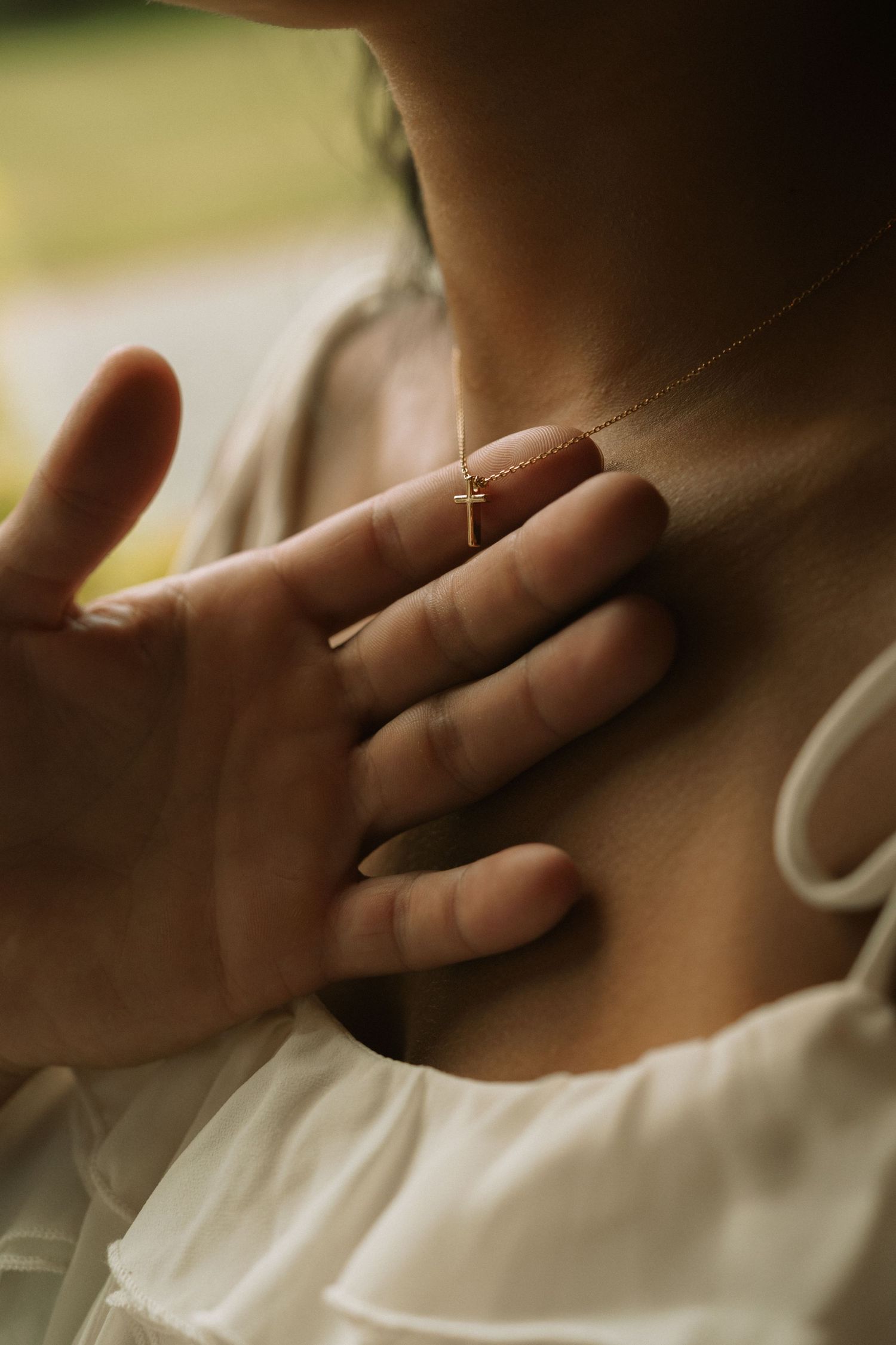 Close-up of a hand touching a delicate gold cross necklace on a woman's chest wearing a white dress.