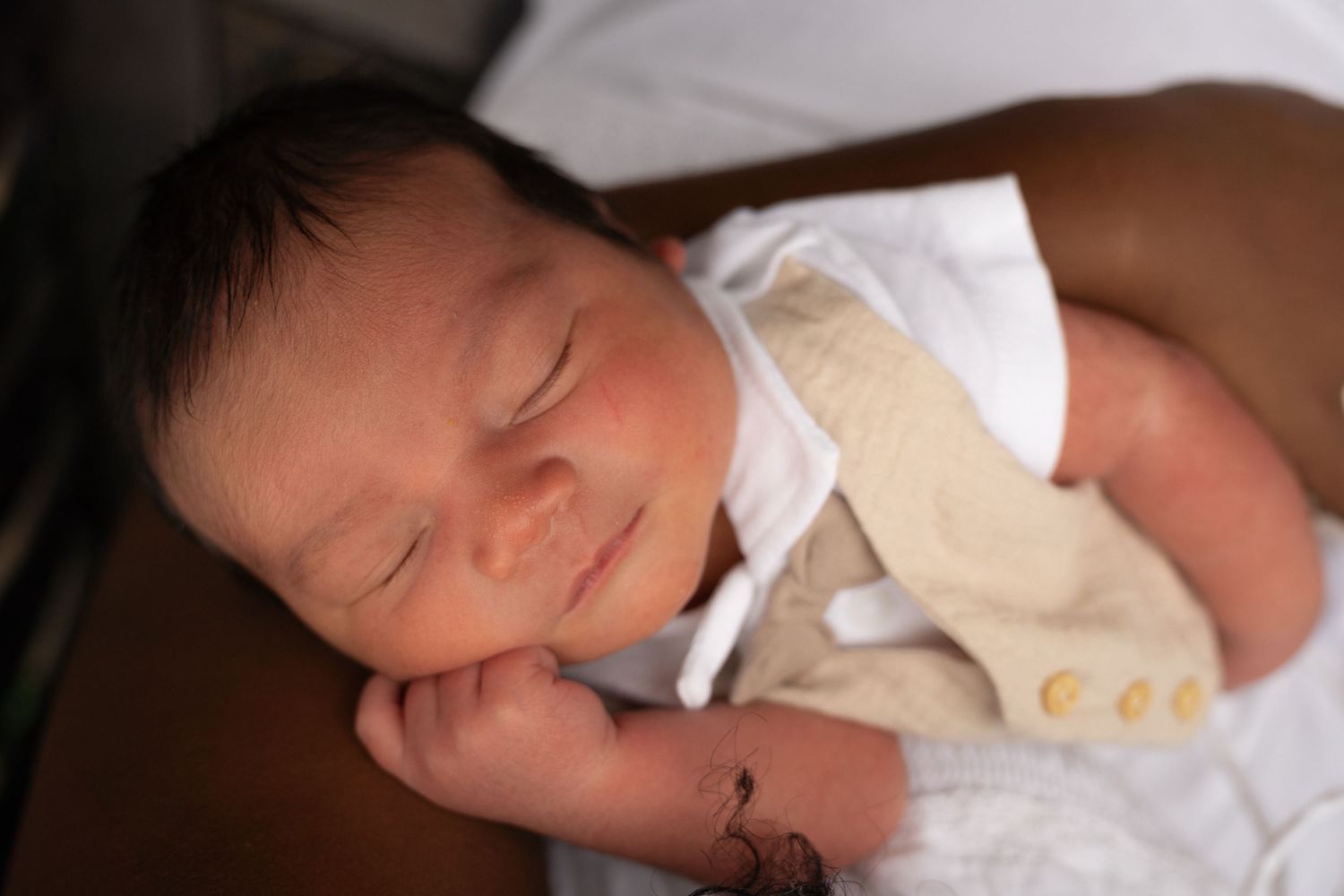 Peaceful newborn baby sleeping with dark hair, dressed in white and beige outfit, smiling softly.