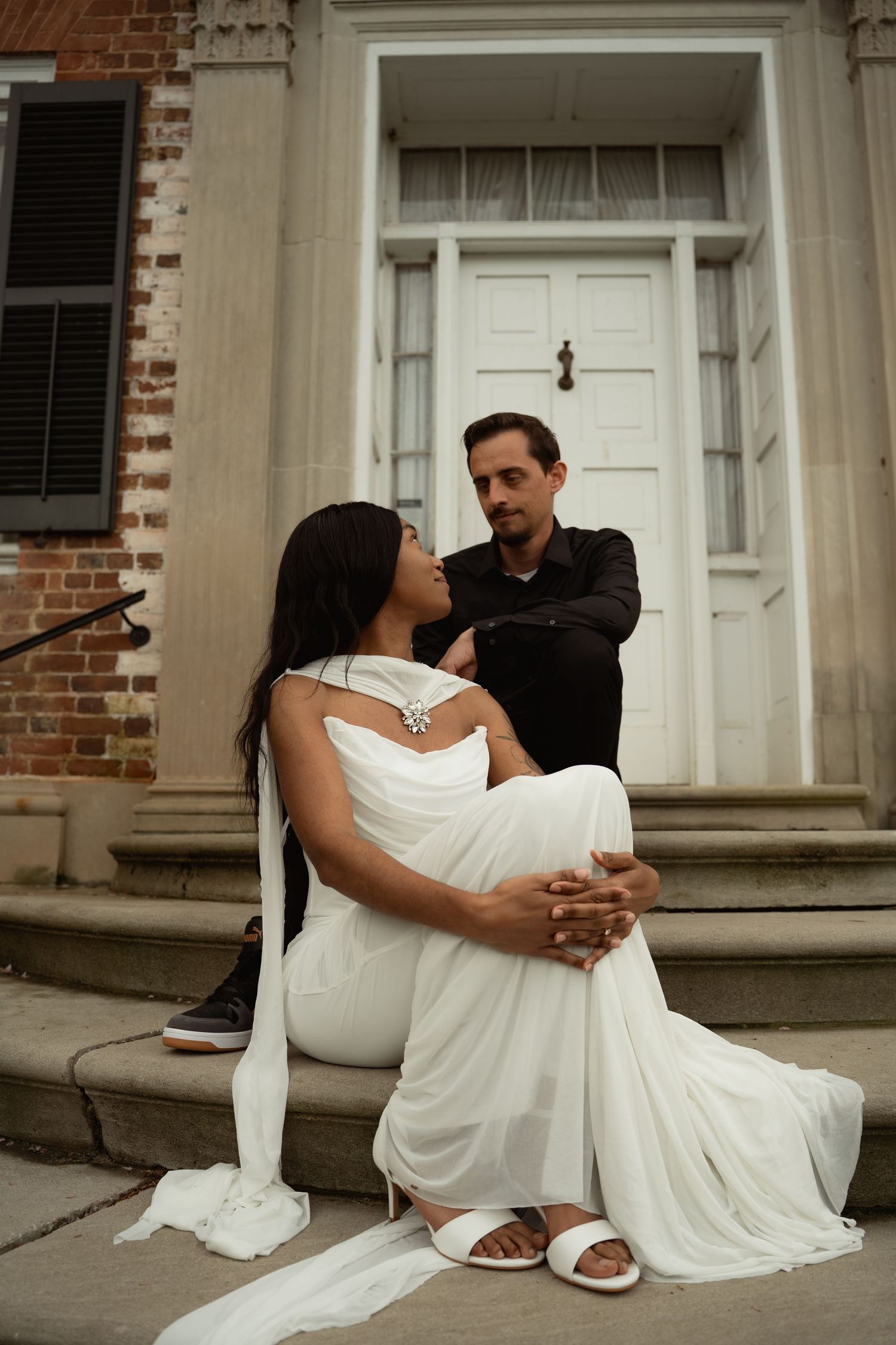 Elegant interracial couple posing on historic brick building steps, woman in flowing white gown. Taken by Row And Co Photography in FXBG VA