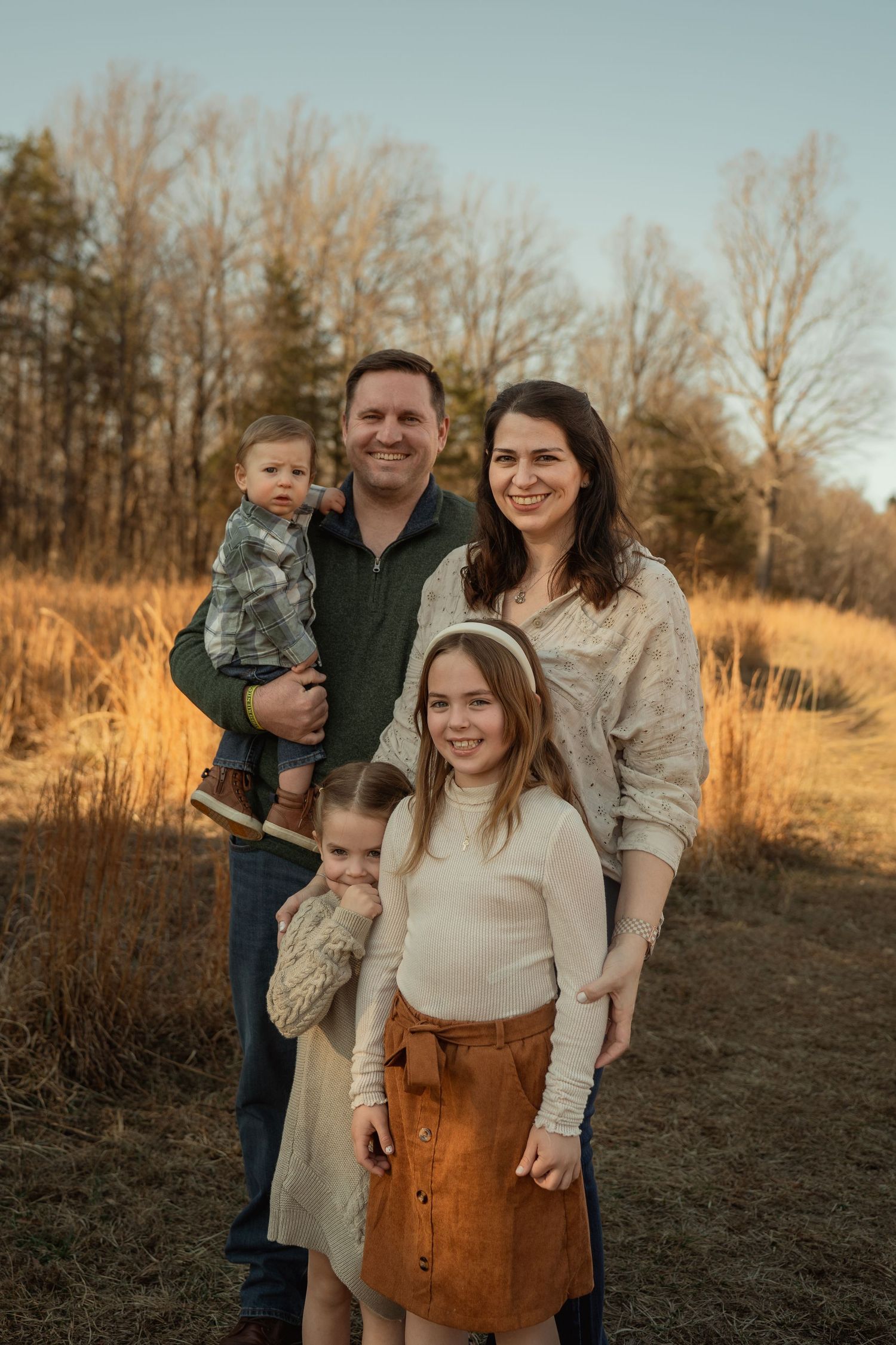 Family of five smiling outdoors in a golden field during autumn sunset, with bare trees in the background. Taken by Row And Co Photography