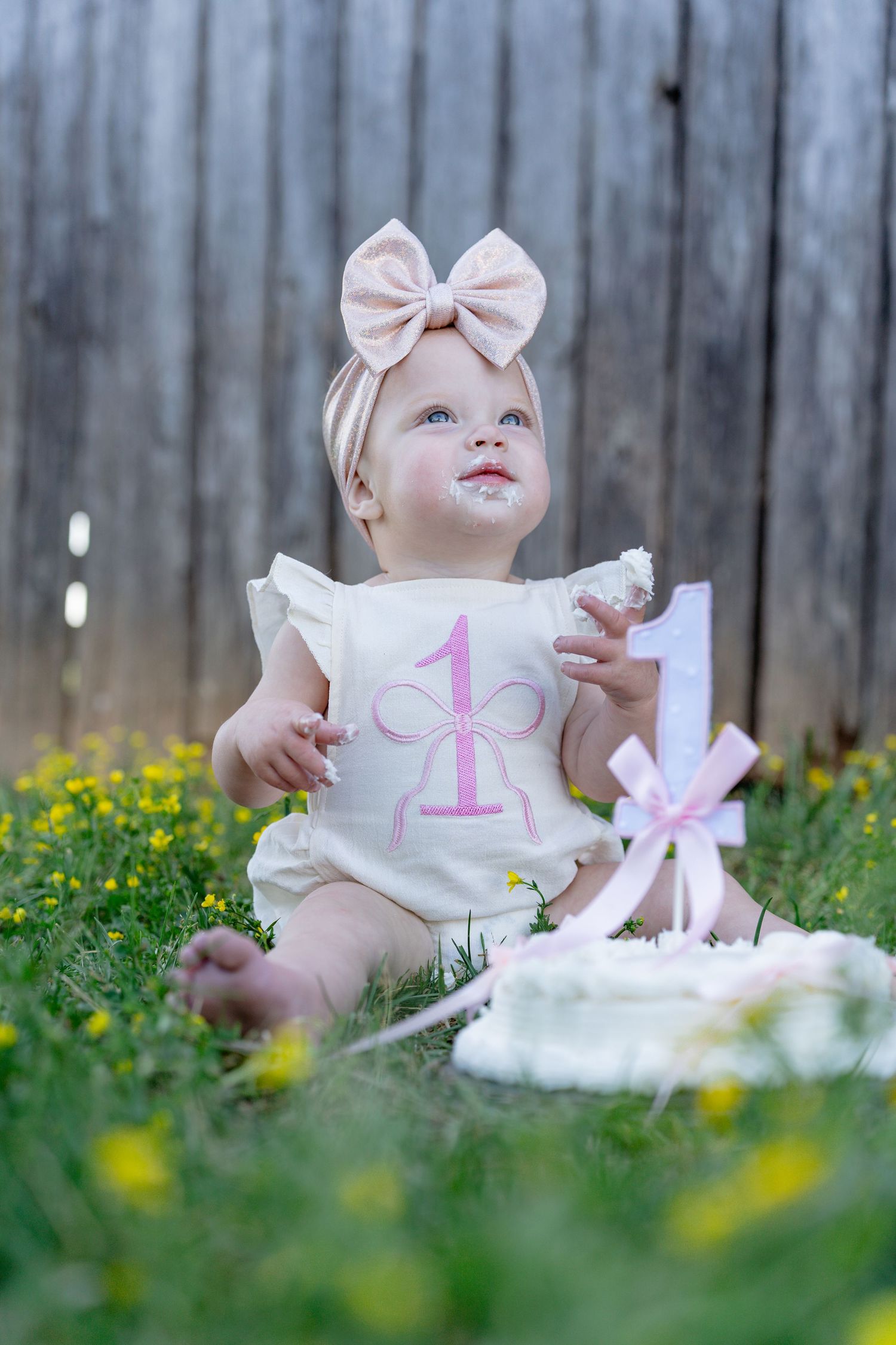 Baby girl in first birthday outfit sitting in grass with smash cake. Taken by Row And Co Photography in FXBG VA