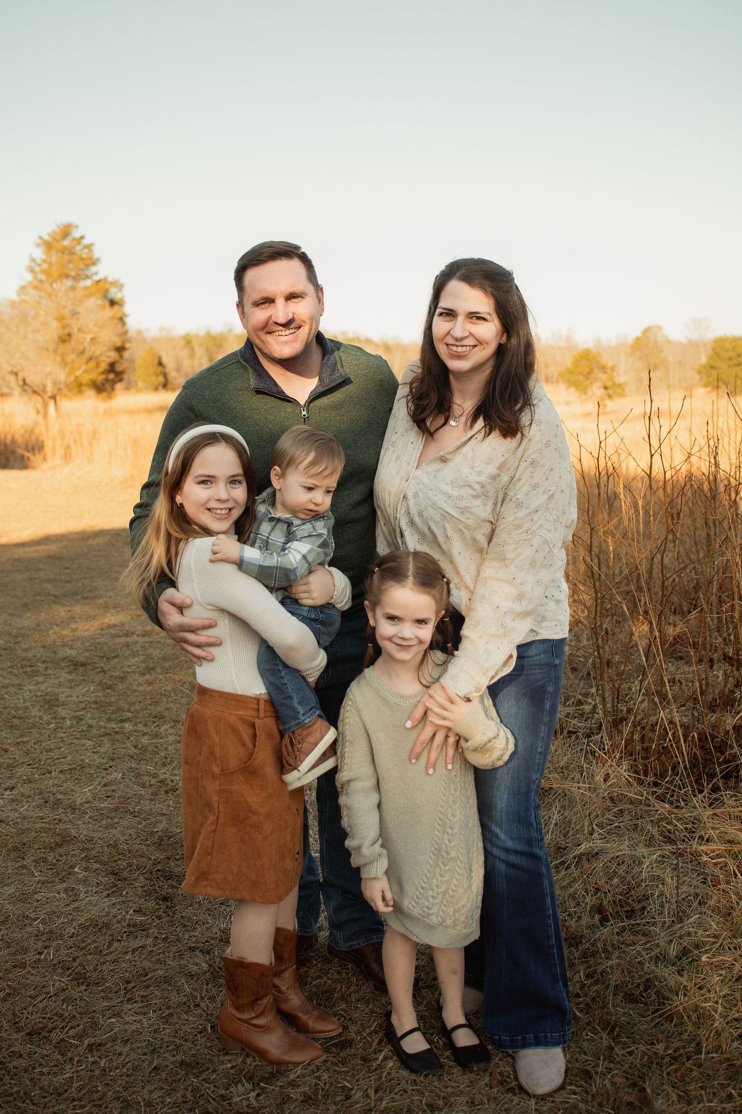 A smiling family of five poses in a golden field at sunset, with two girls and a toddler boy. Taken by Row And Co Photography in FXBG VA