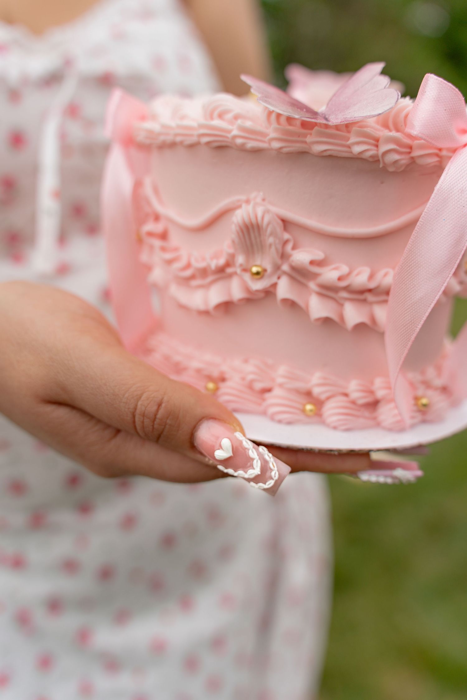 Woman in floral dress holding an ornate pink cake with ruffles, bows, and gold pearl decorations outdoors. Taken by Row And Co Photography