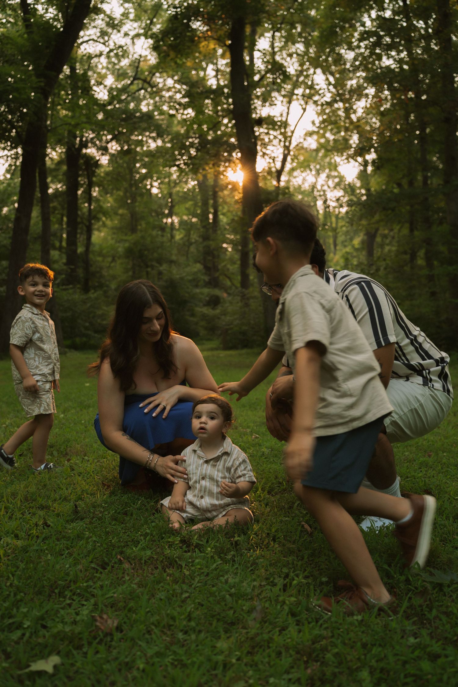 A mother sits on grass with her baby while children play around her at golden hour in a lush green forest.