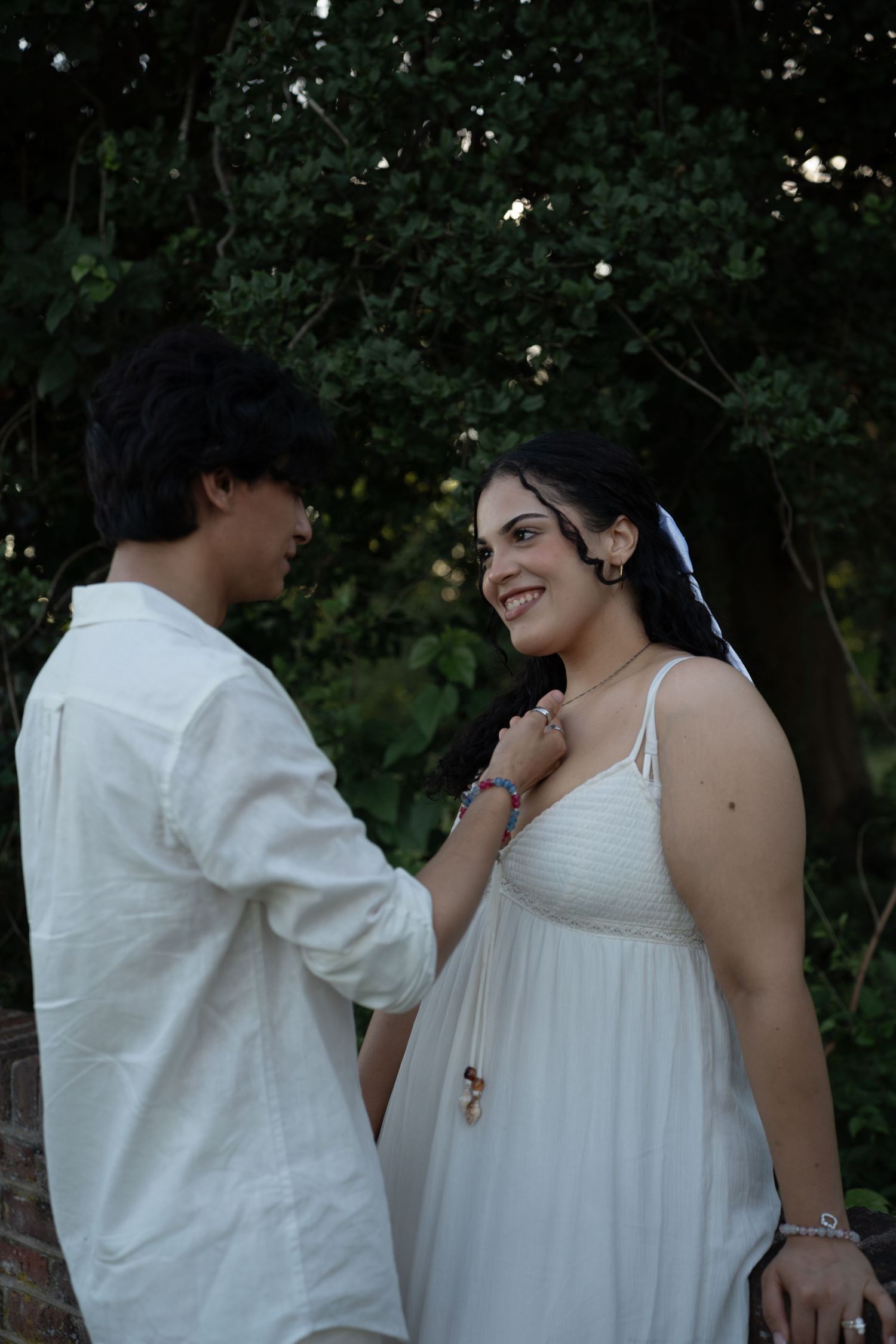 A young couple in white attire share a romantic moment outdoors, surrounded by lush green trees.