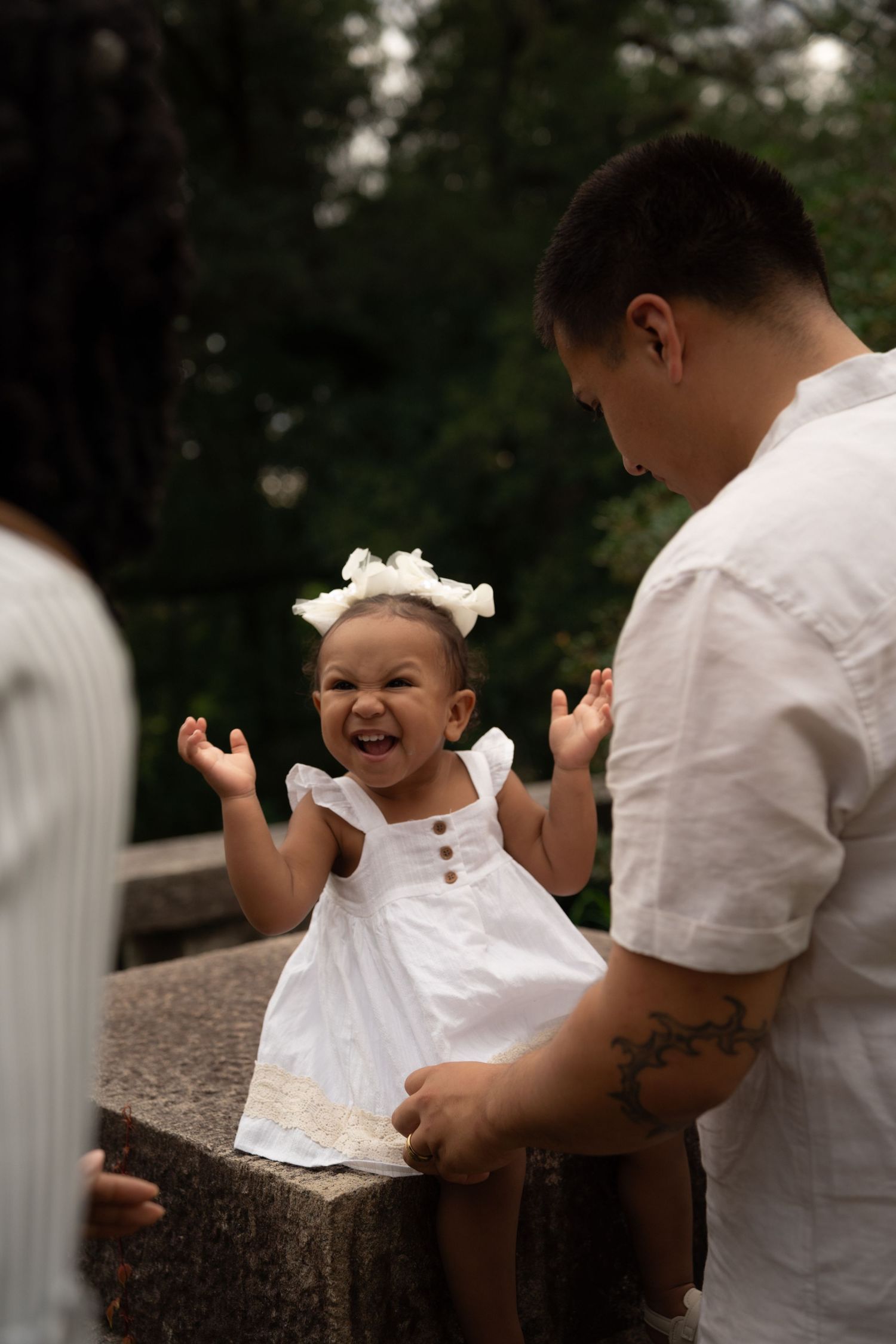 Laughing baby girl in white dress and bow headband sits on stone ledge with tattooed father outdoors.