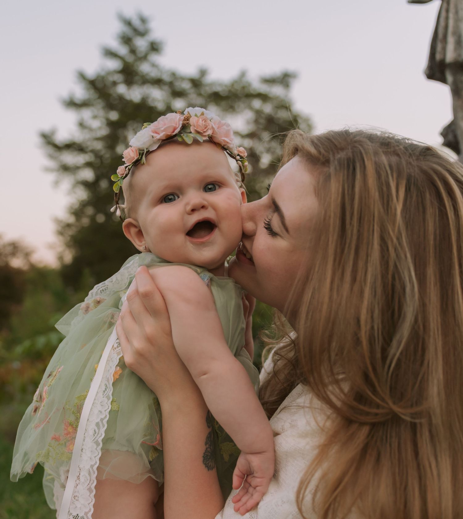 A woman kisses a smiling baby girl wearing a floral crown and green dress outdoors.