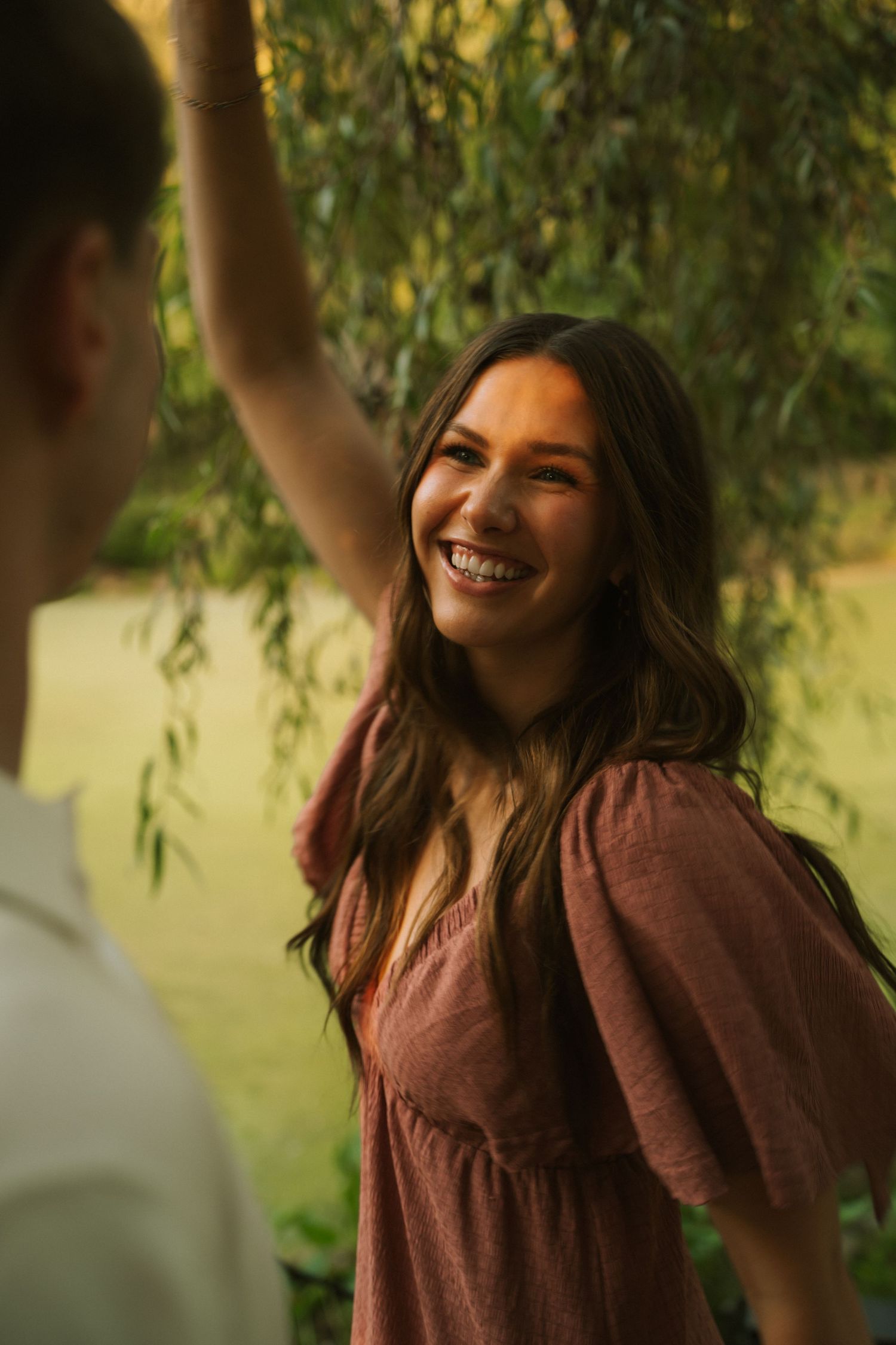 A smiling young woman in a pink dress laughs joyfully outdoors near a willow tree with a green pond backdrop.