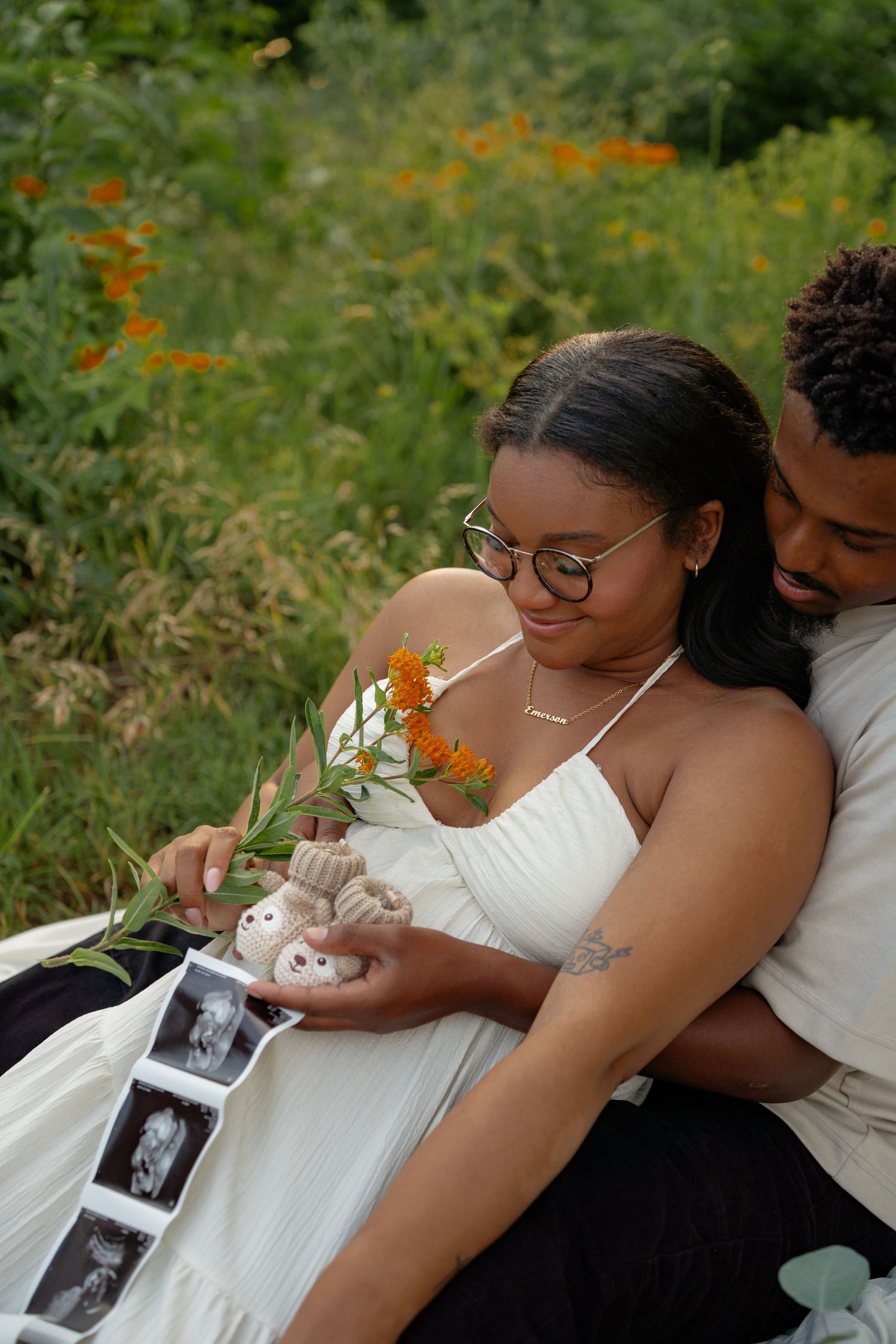 Expecting couple sits in a wildflower field holding ultrasound photos and tiny baby booties together.