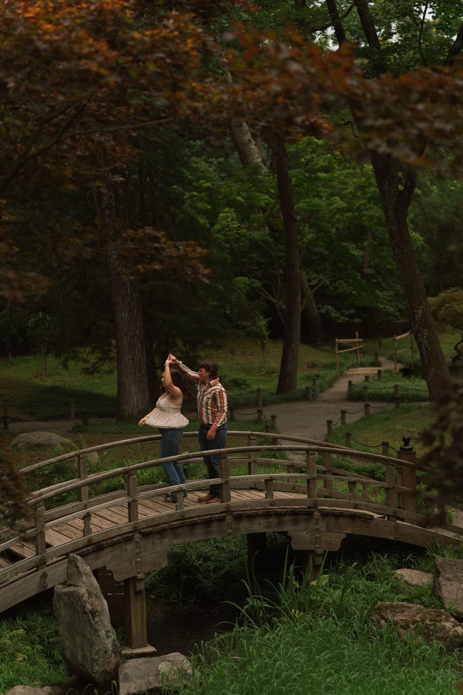 Couple dancing on a wooden arched bridge surrounded by lush green trees in a serene park setting.
