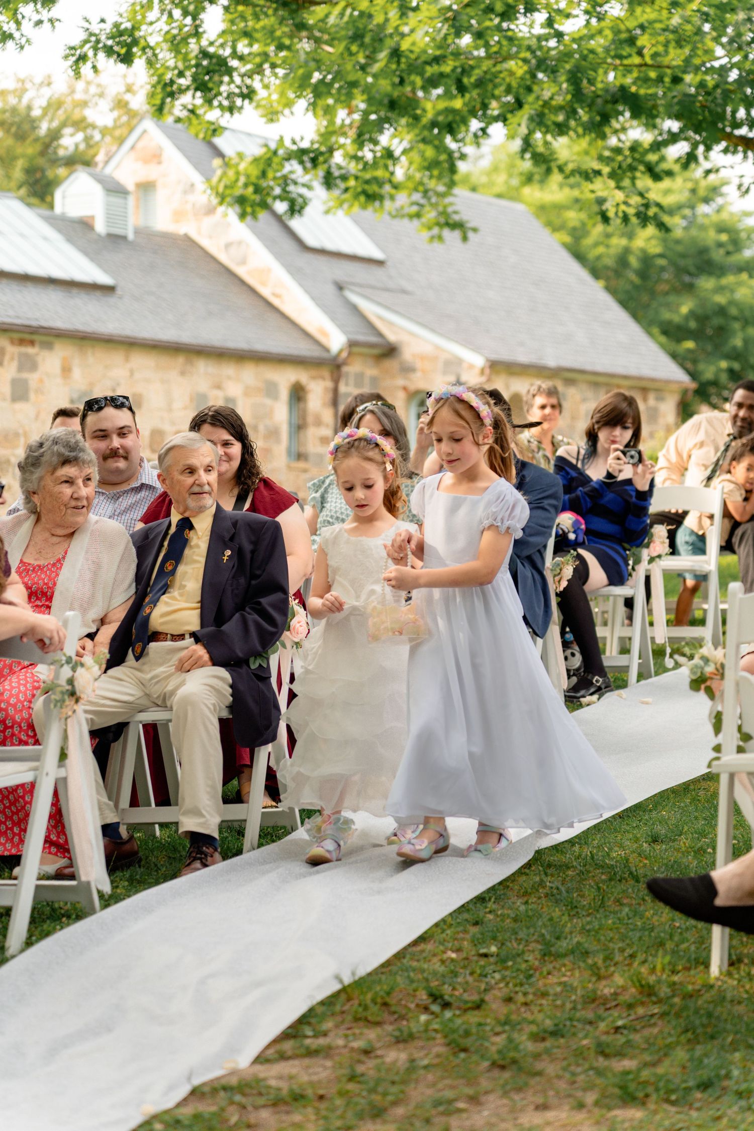 Two flower girls in white dresses walk down the aisle at an outdoor wedding ceremony with guests seated on white chairs.