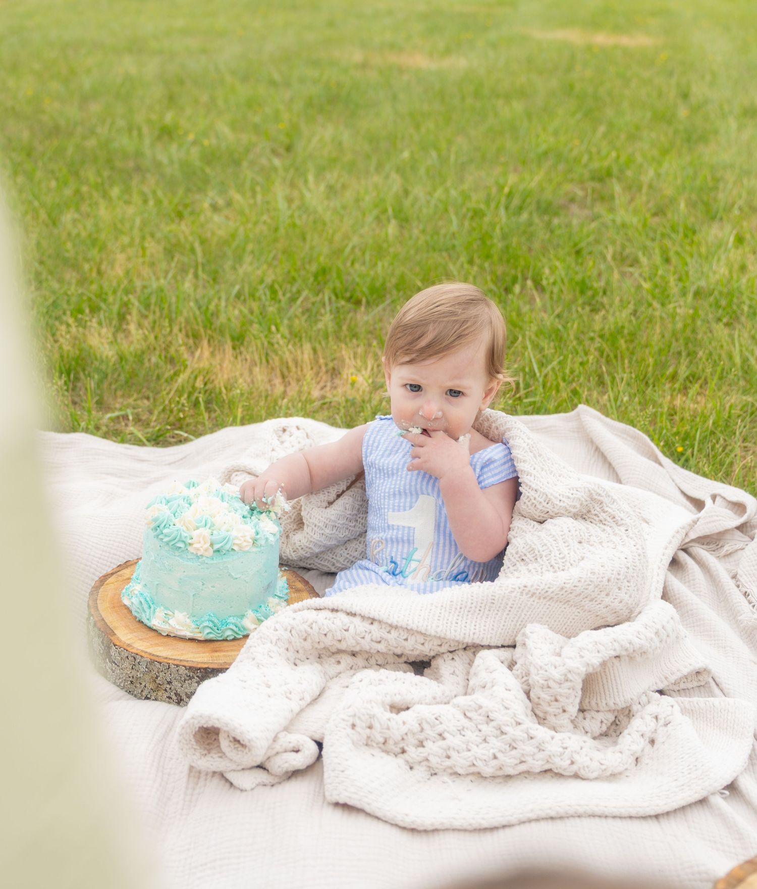 Baby celebrating first birthday with a teal smash cake on a blanket in a grassy field. Taken by Row And Co Photography in FXBG VA