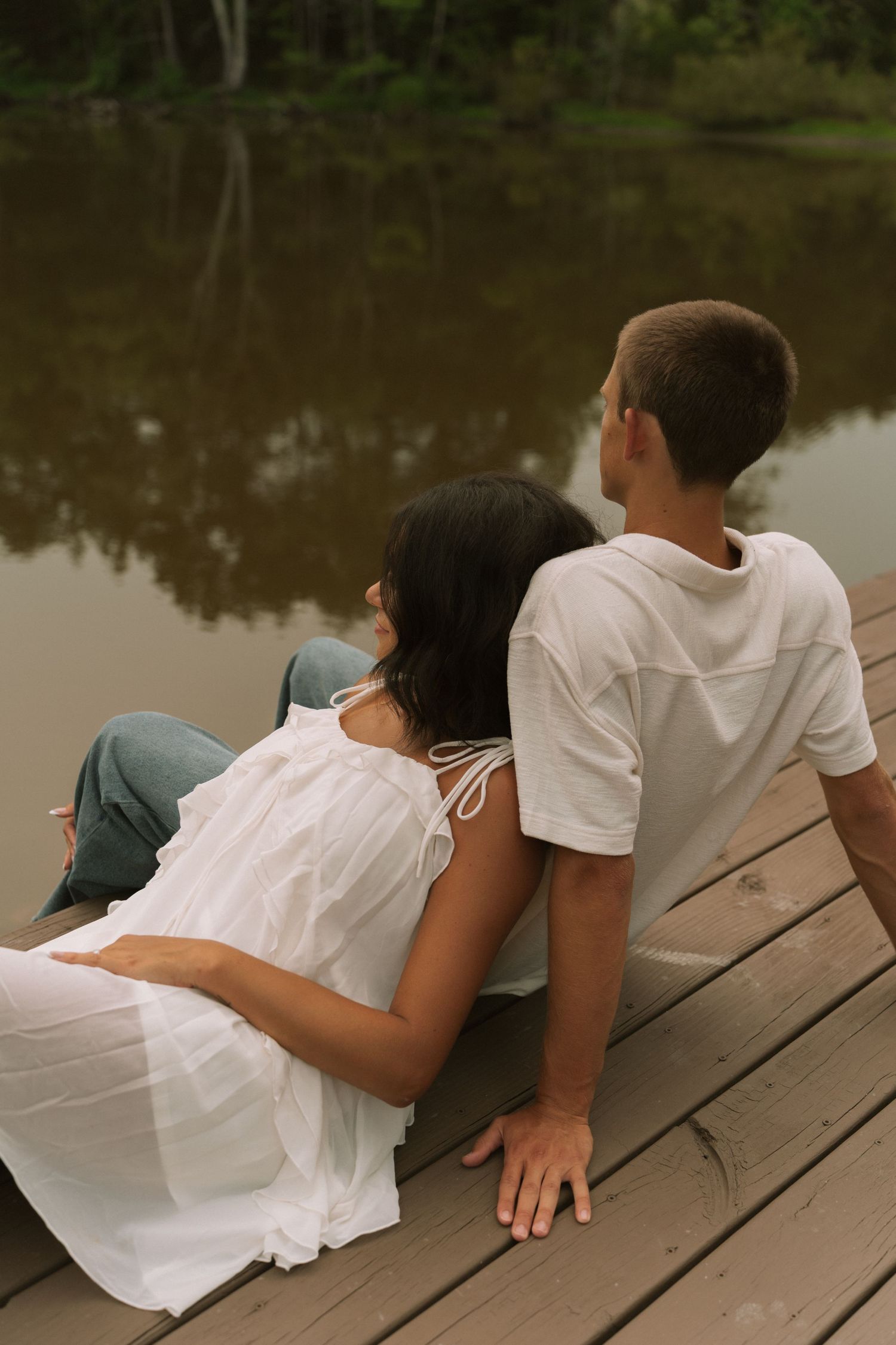 A young couple sits together on a wooden dock, gazing at a calm reflective lake surrounded by lush green trees.