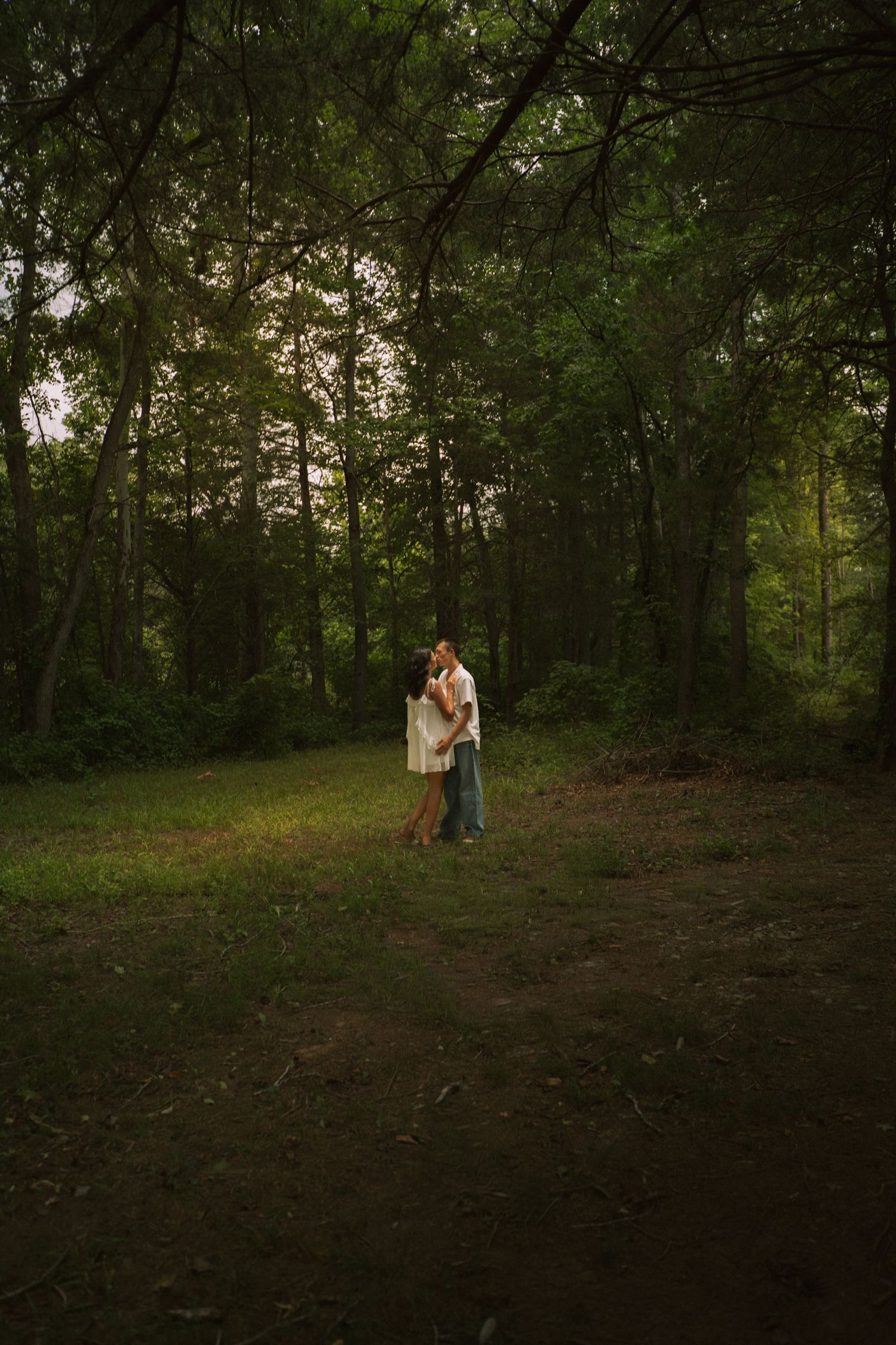 Couple sharing a romantic embrace in a lush green forest clearing at dusk.