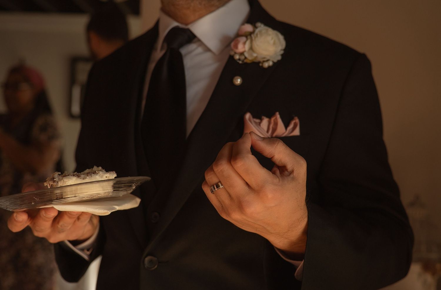Groom in black suit with floral boutonniere holds a plate of wedding cake at the reception.