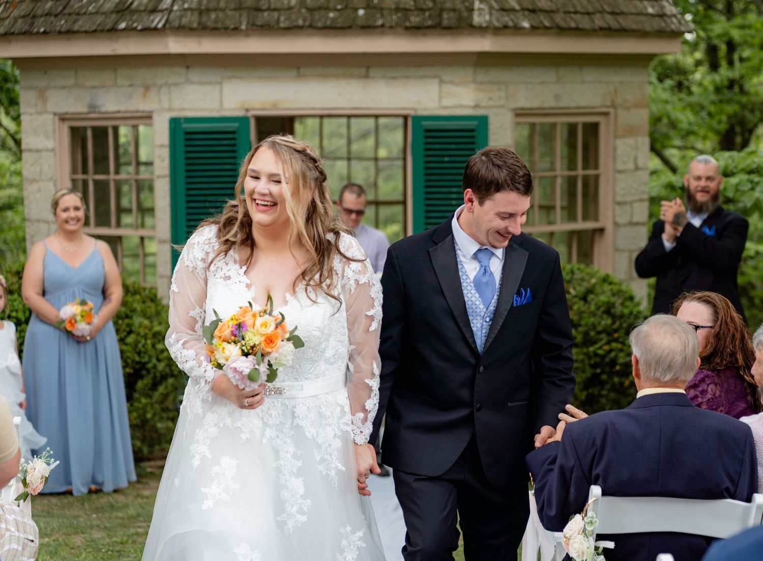 Happy bride and groom walk down the aisle at an outdoor garden wedding ceremony with guests applauding.