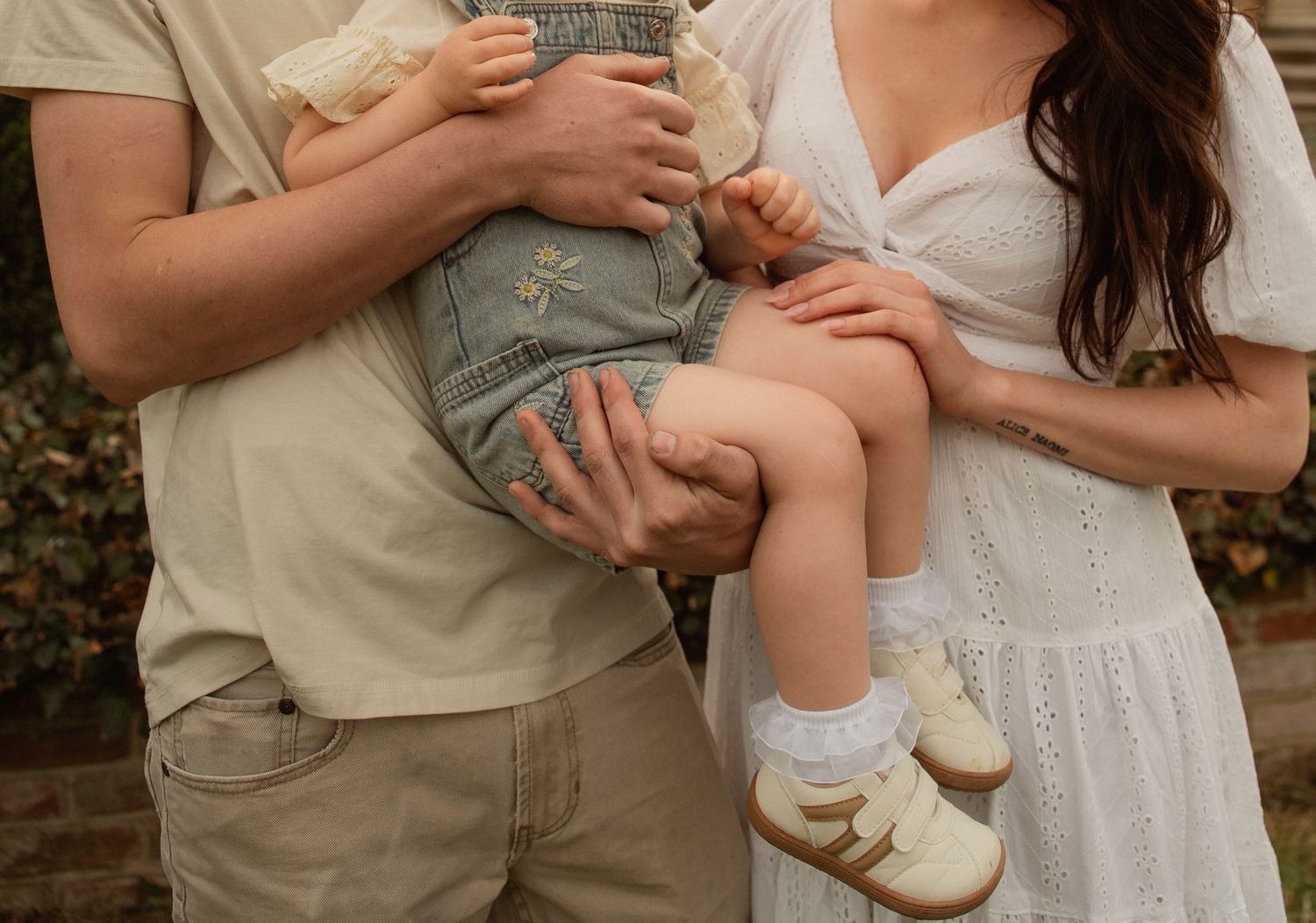 Parents in neutral tones hold their baby dressed in denim overalls and cream shoes outdoors.Taken by Row And Co Photography in FXBG VA