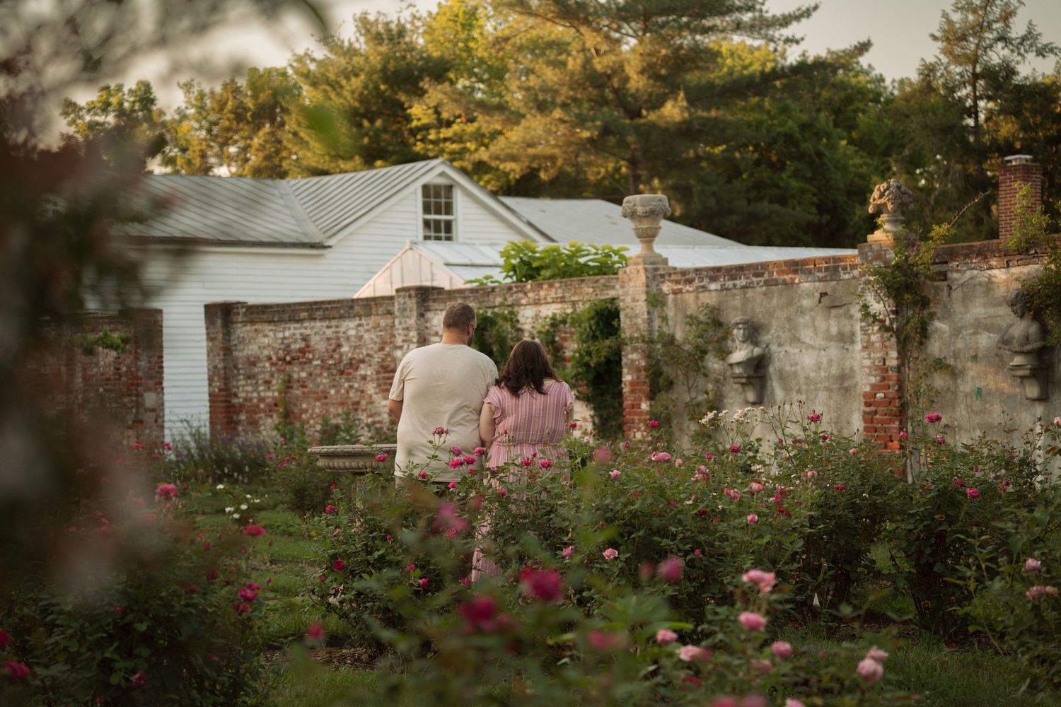 A couple walks hand in hand through a lush garden with blooming flowers toward a white cottage. Taken by Row And Co Photography in FXBG VA