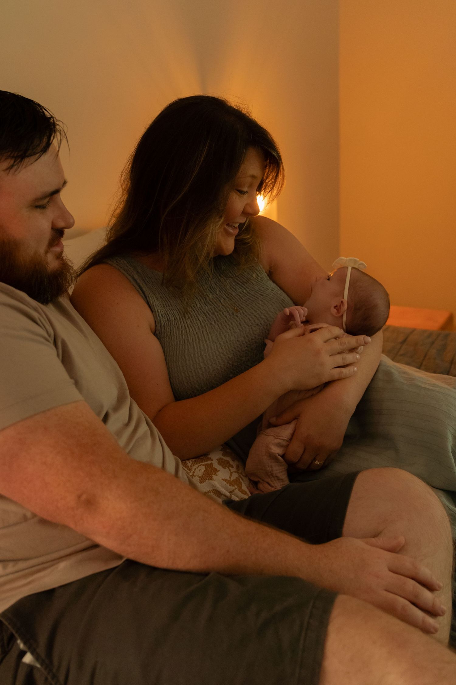 A couple sits together in warm light, smiling at their newborn baby, sharing a tender family moment.