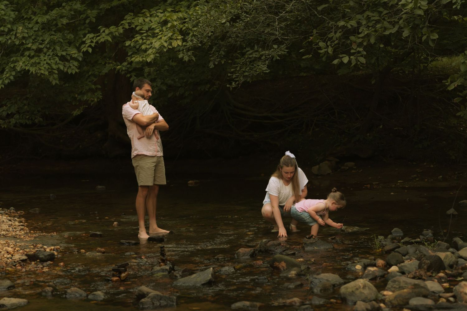 Family wading in a rocky shallow creek surrounded by lush green trees, with children playing near the water.