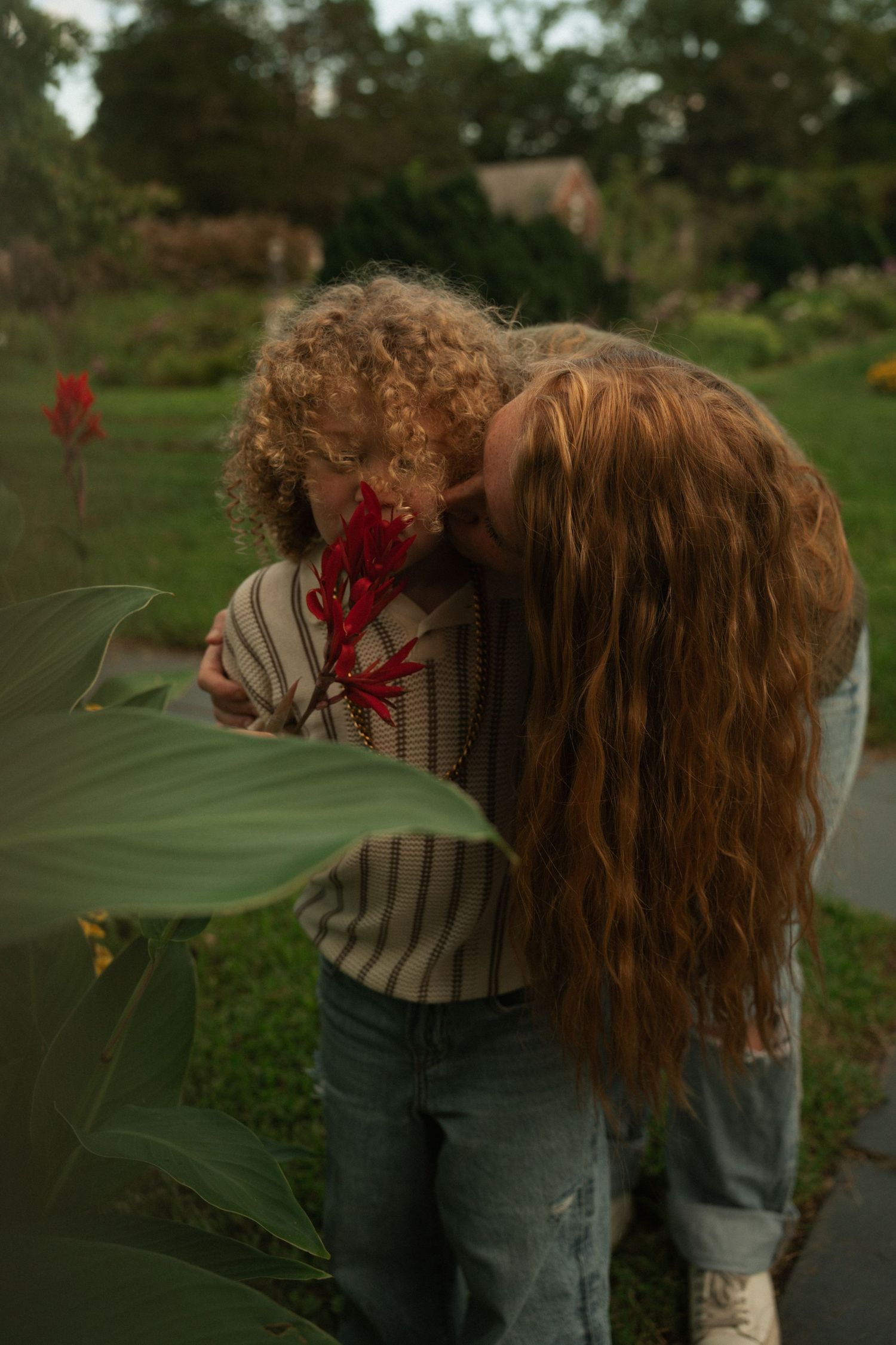 Two curly-haired people smelling a red flower together in a lush green garden setting.