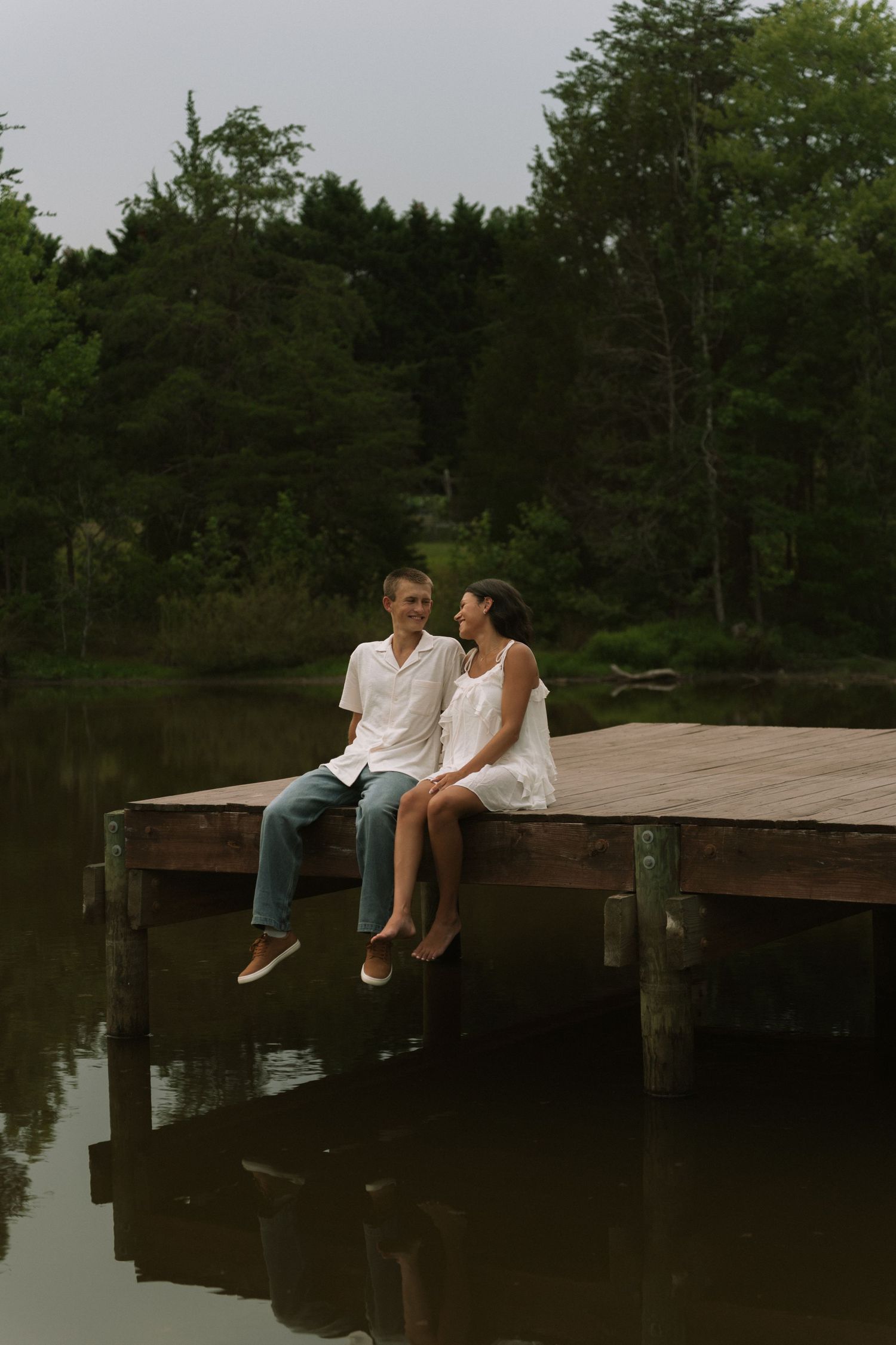 Couple sitting on a wooden dock over a calm lake, surrounded by lush green trees, sharing a romantic moment.
