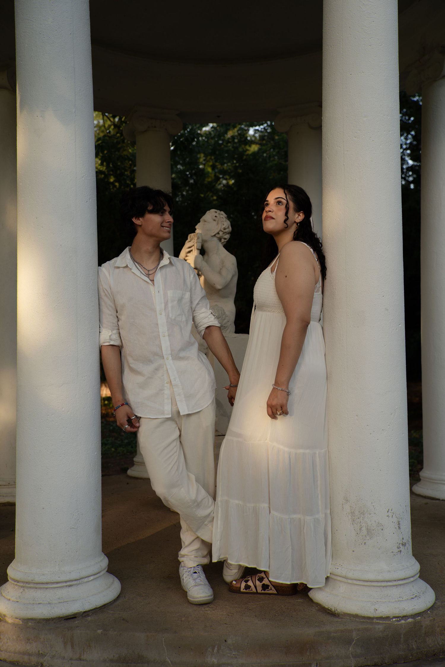 Couple in white outfits holding hands between classical columns with a statue backdrop in a garden setting.