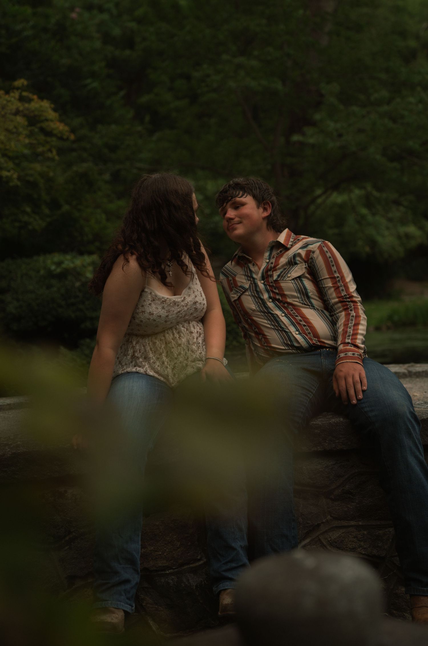A young couple sits together on a stone ledge in a lush green park, gazing at each other romantically.