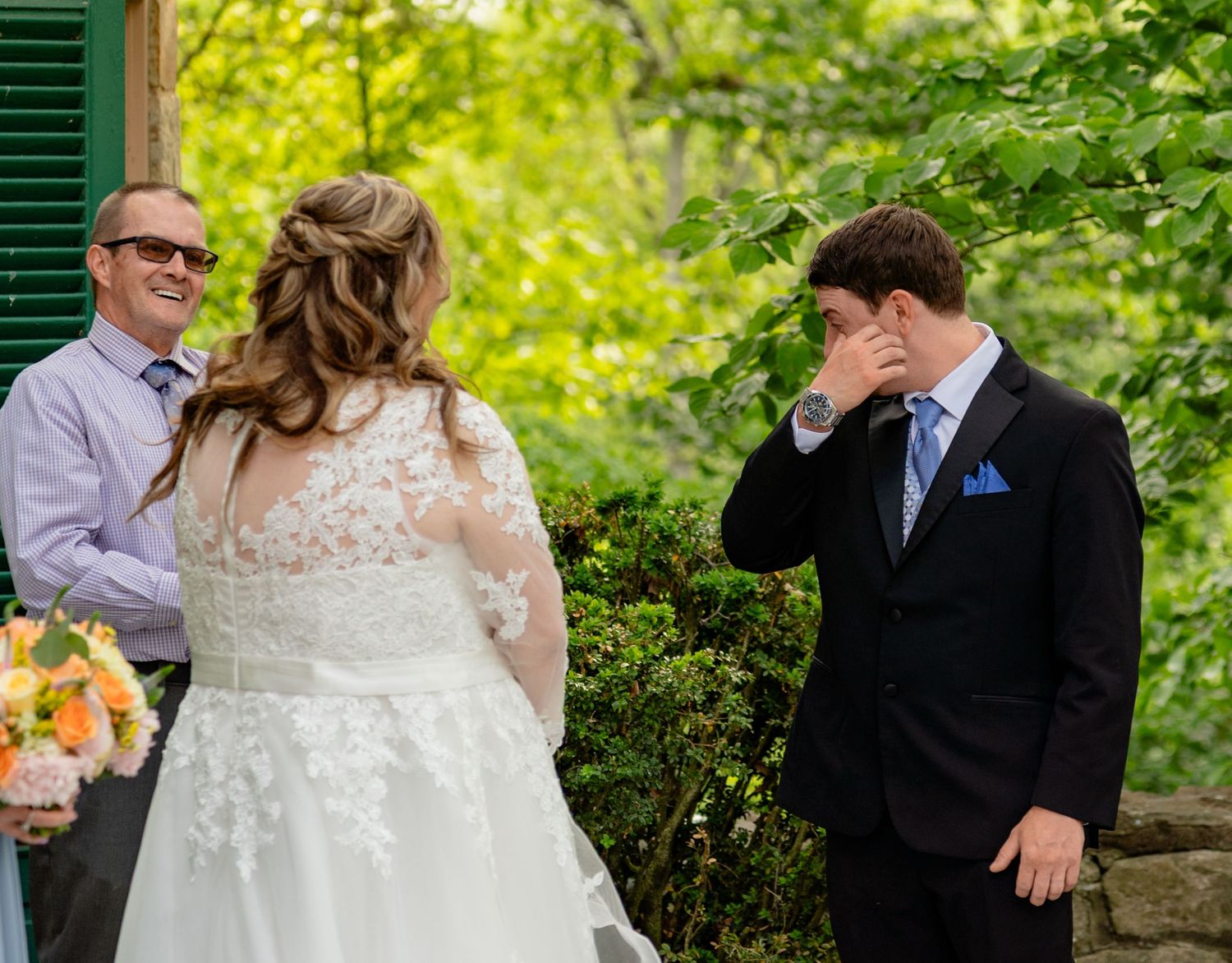 Emotional groom wiping tears as bride approaches during outdoor wedding ceremony with greenery backdrop.