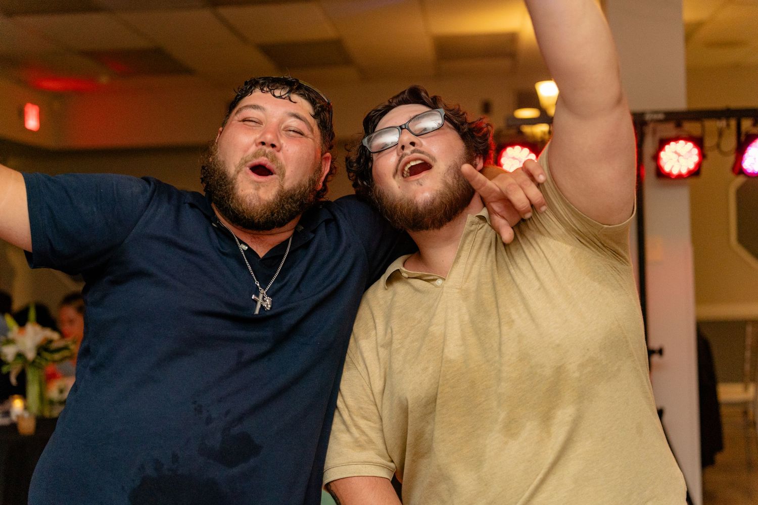 Two young men celebrating joyfully at a party, one pointing up while posing together in a banquet hall.