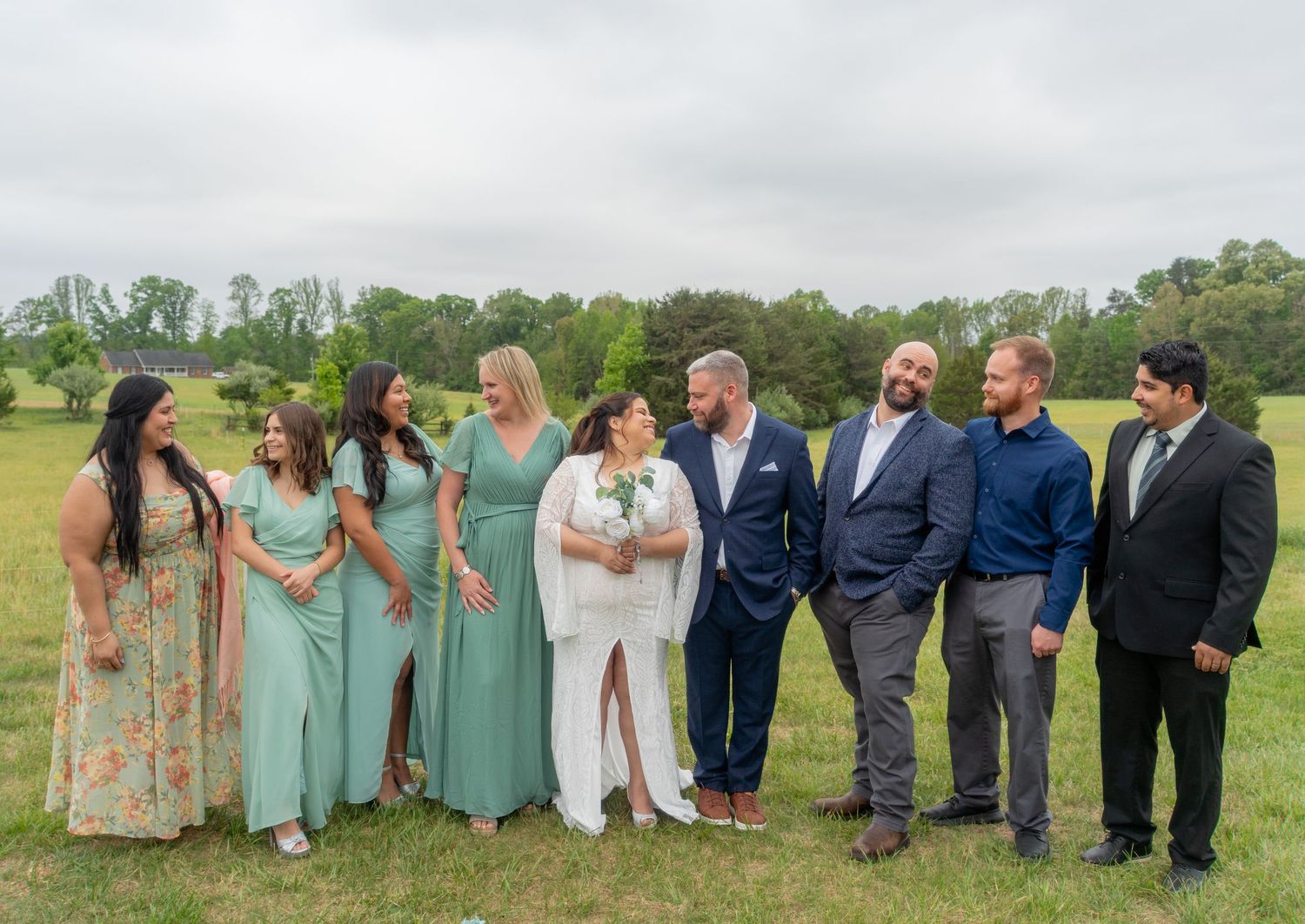 Wedding party poses outdoors on green lawn, bride in white, bridesmaids in sage green dresses, groomsmen in navy suits.