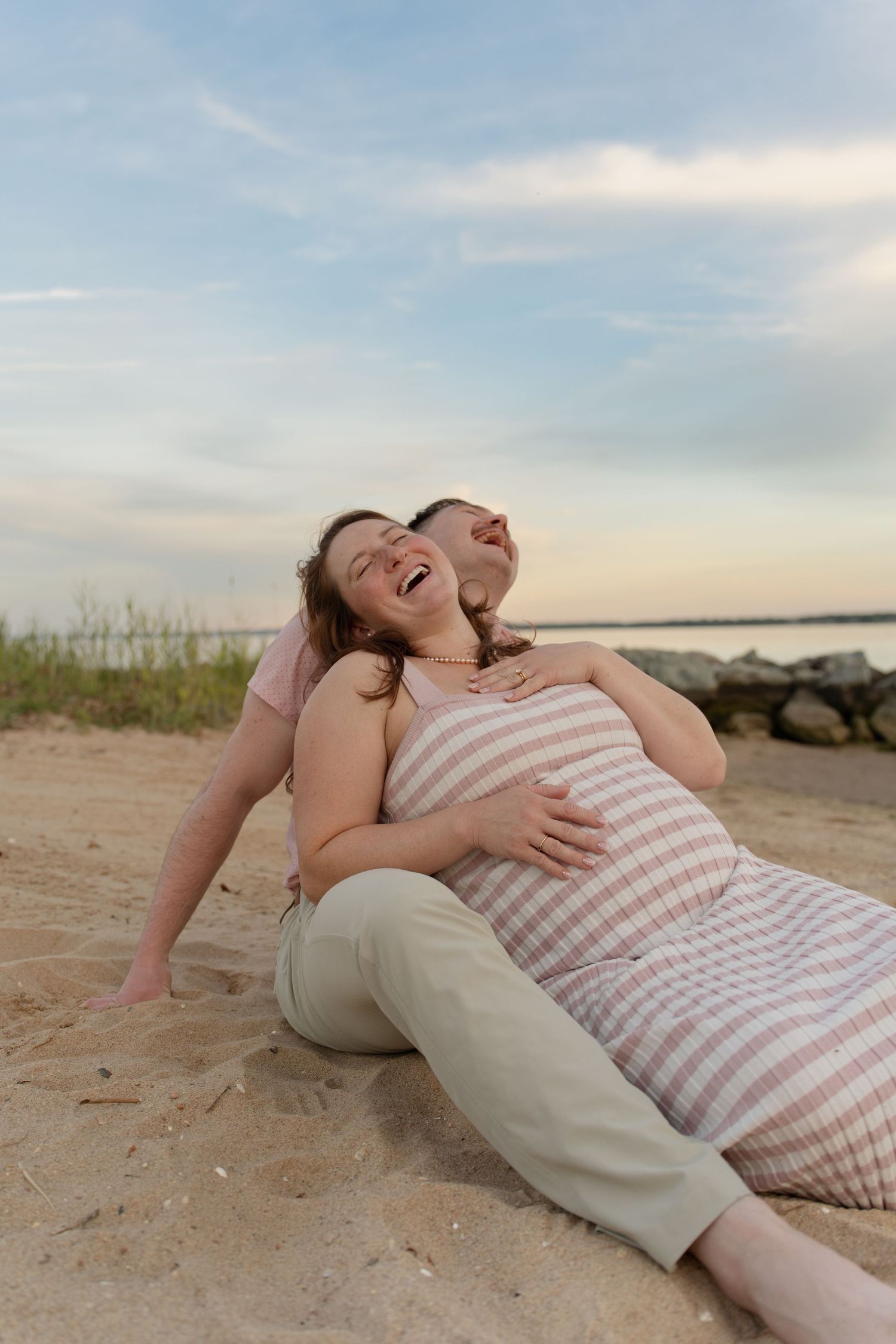 Laughing pregnant woman sits on sandy beach leaning against partner who embraces her baby bump at sunset. Taken by Row And Co Photography