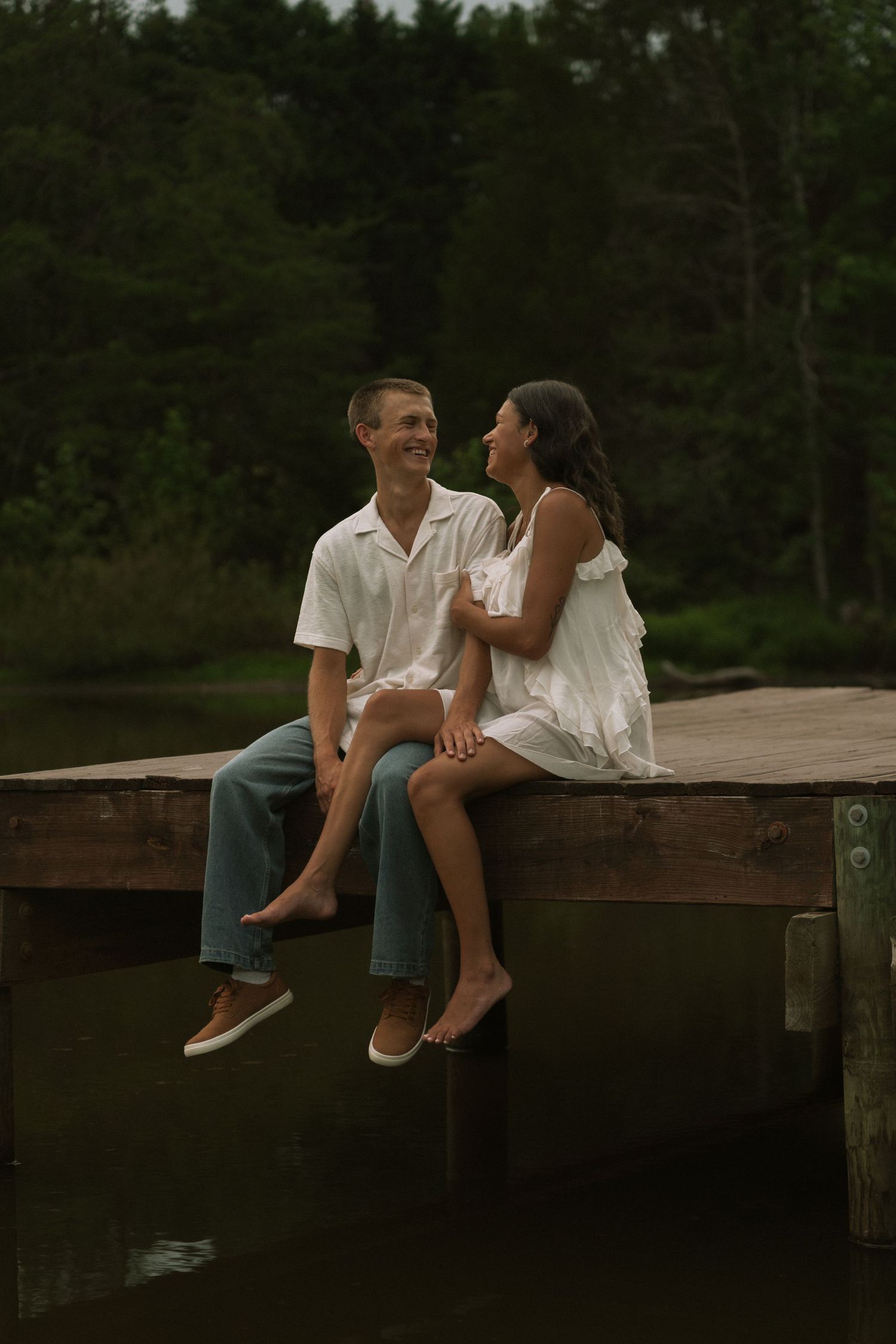A couple sits on a wooden dock by a lake, smiling at each other, dressed in white and casual attire.