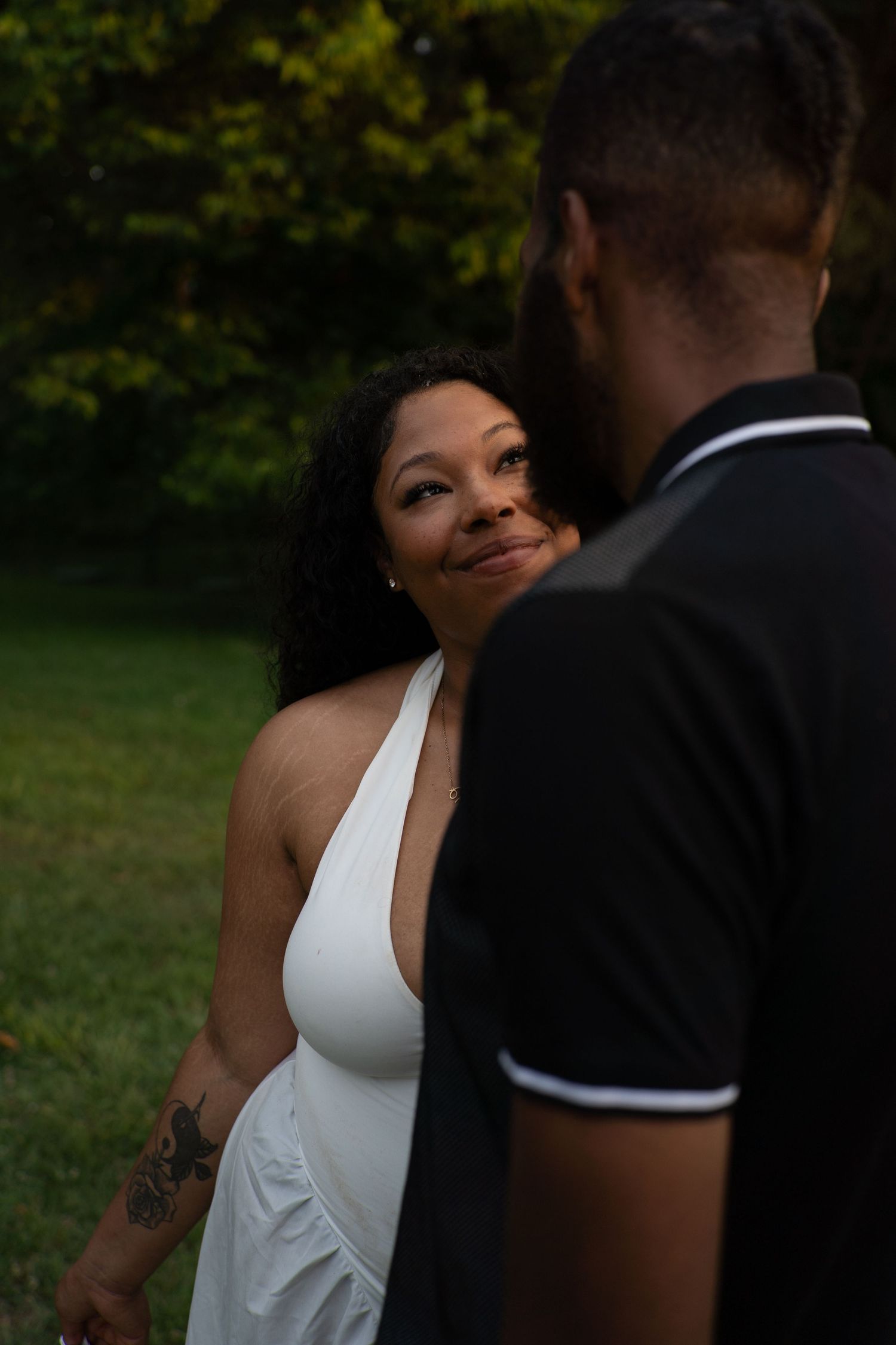 A woman in a white halter dress smiles lovingly at a man in a black polo shirt outdoors.
