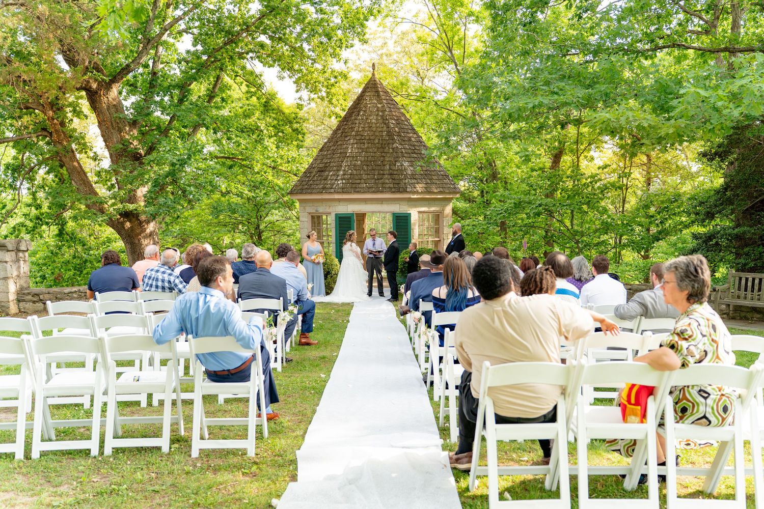 Outdoor wedding ceremony with white chairs on a green lawn leading to a small chapel surrounded by lush trees.