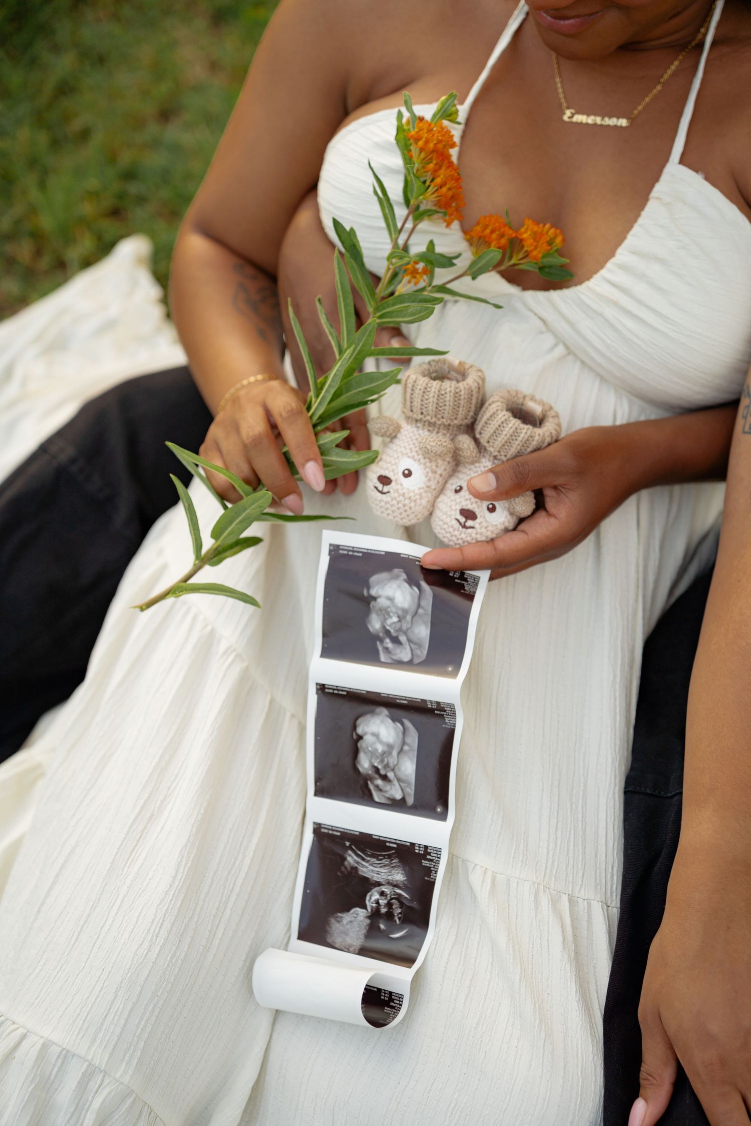 Pregnant woman in white dress holding ultrasound photos, tiny knit baby booties, and orange wildflowers outdoors.