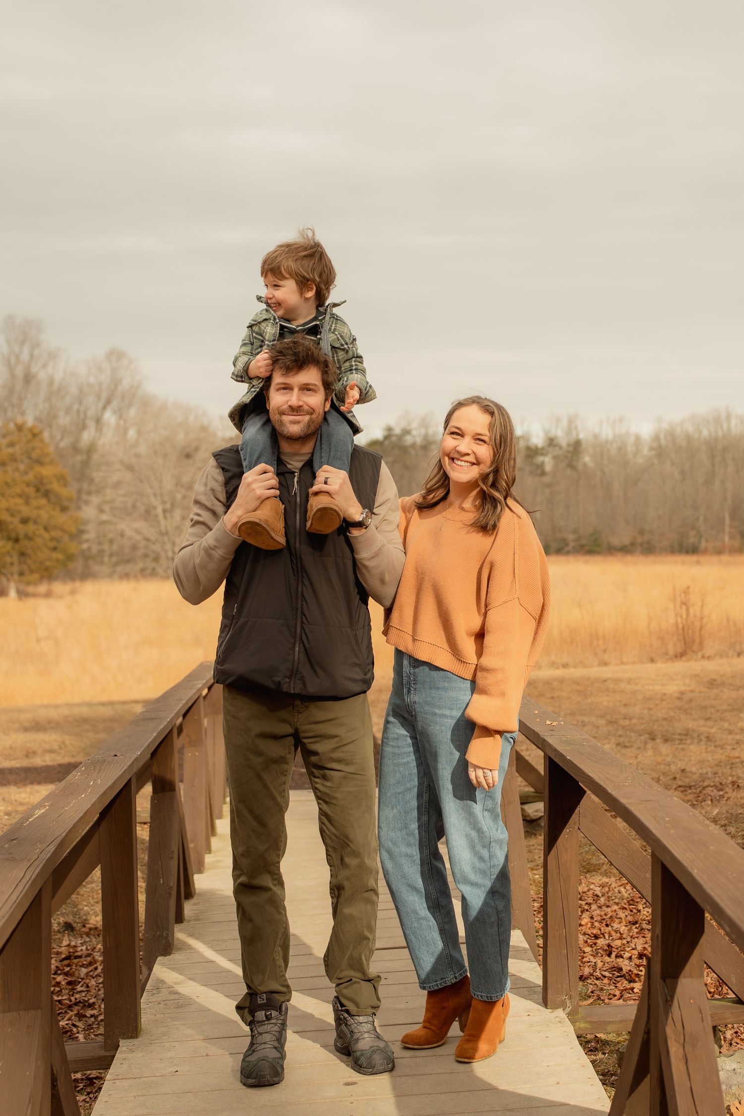 Family of three posing on a wooden bridge in autumn, father carrying toddler on shoulders beside smiling mother in orange sweater.